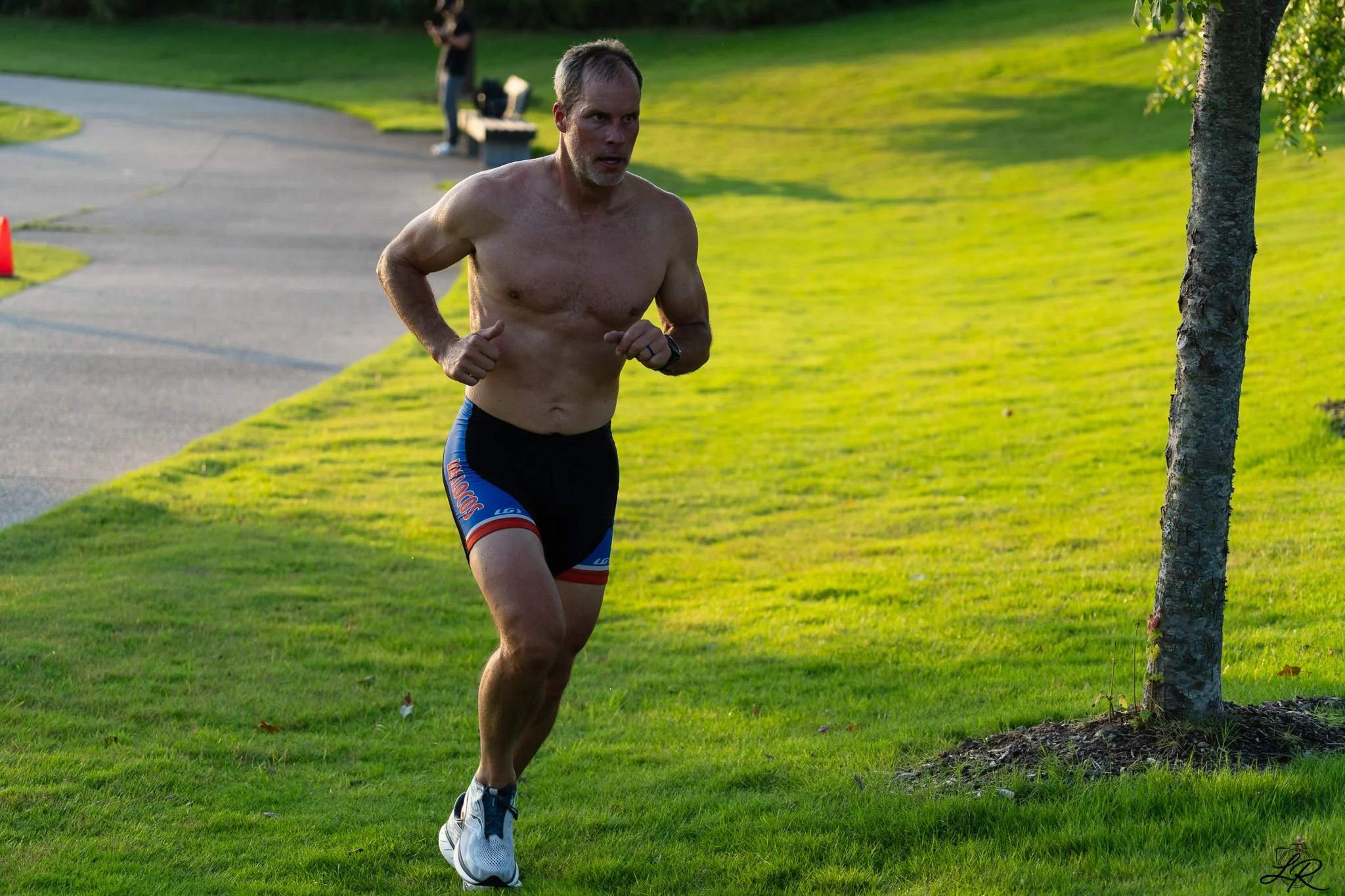 A shirtless man running outdoors on a grassy path near a tree in a park during late afternoon or early evening.