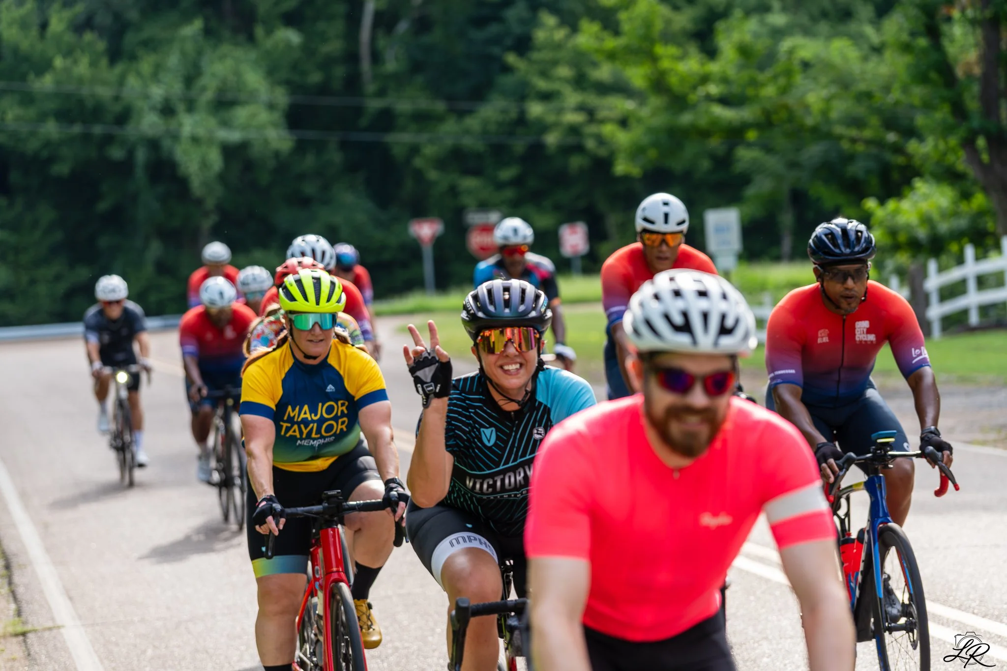 Group of cyclists riding on a road surrounded by trees, some wearing helmets and sunglasses, with a woman in the front smiling and making a peace sign.