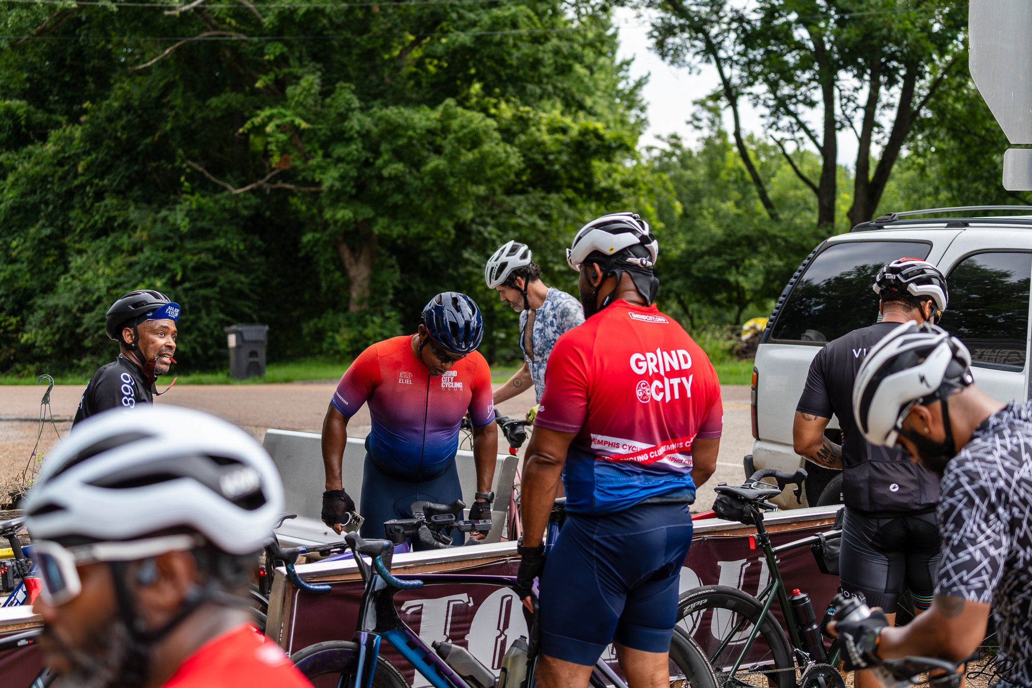 Group of cyclists with helmets gathered near a vehicle on a street, with lush green trees in the background.