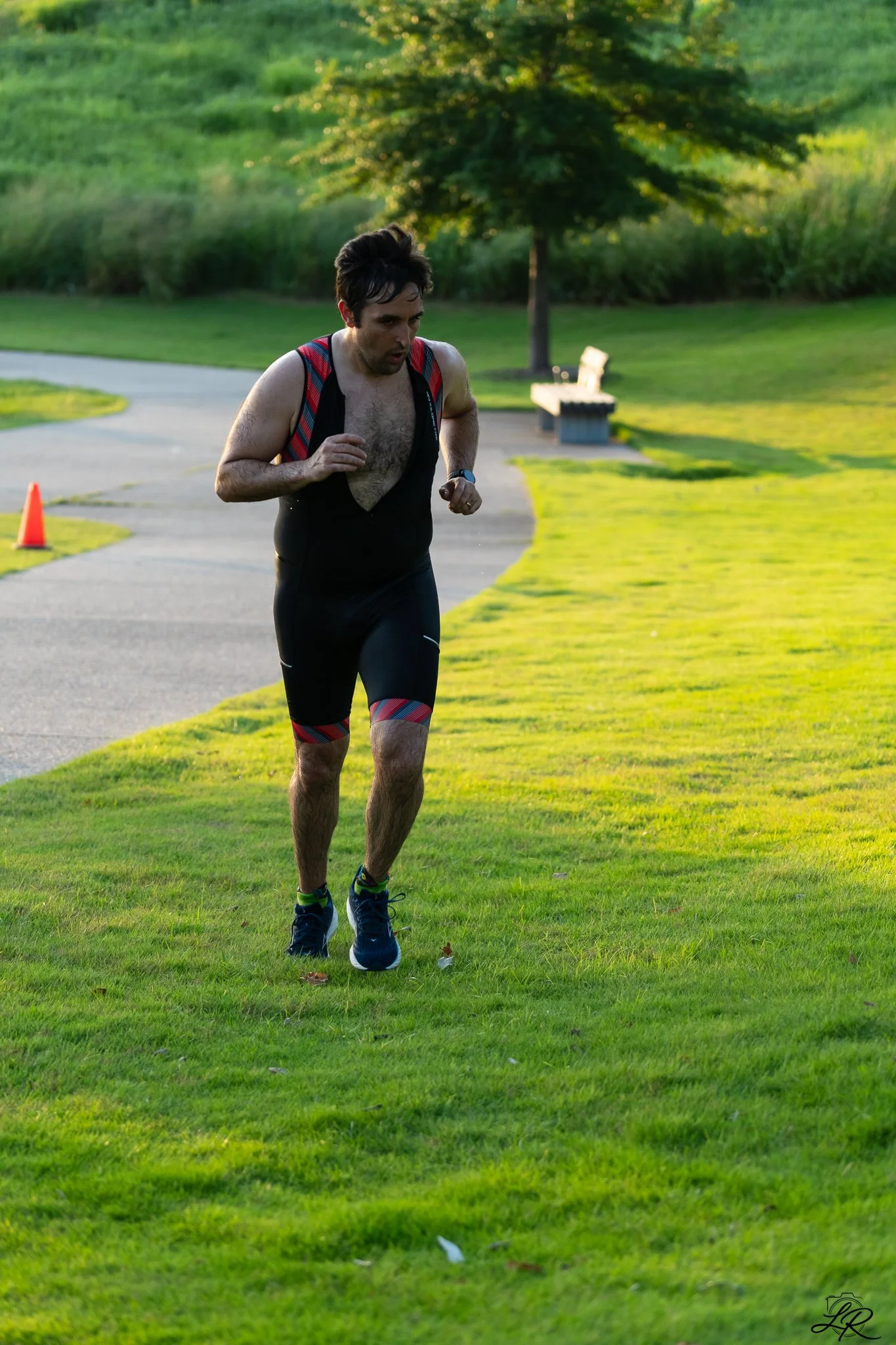 A man jogging outdoors on a grassy park path during sunset, wearing a black and red athletic outfit, with trees and a bench in the background.