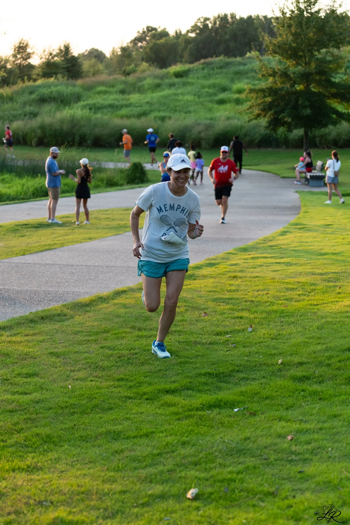 People participating in a running event in a park during sunset, with some walking, sitting, and chatting on benches, and lush green trees and hills in the background.