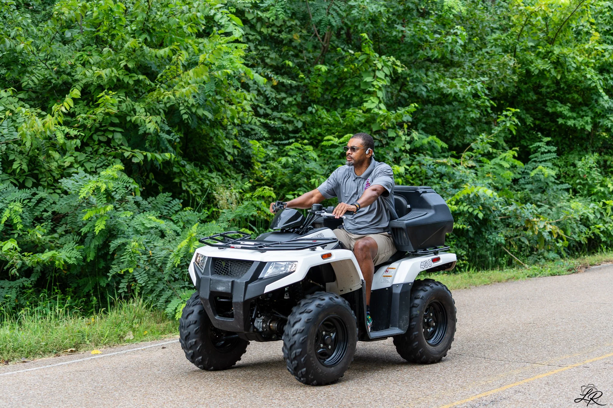 A man wearing sunglasses and earbuds riding a four-wheeler ATV on a paved road, surrounded by lush green trees and bushes.