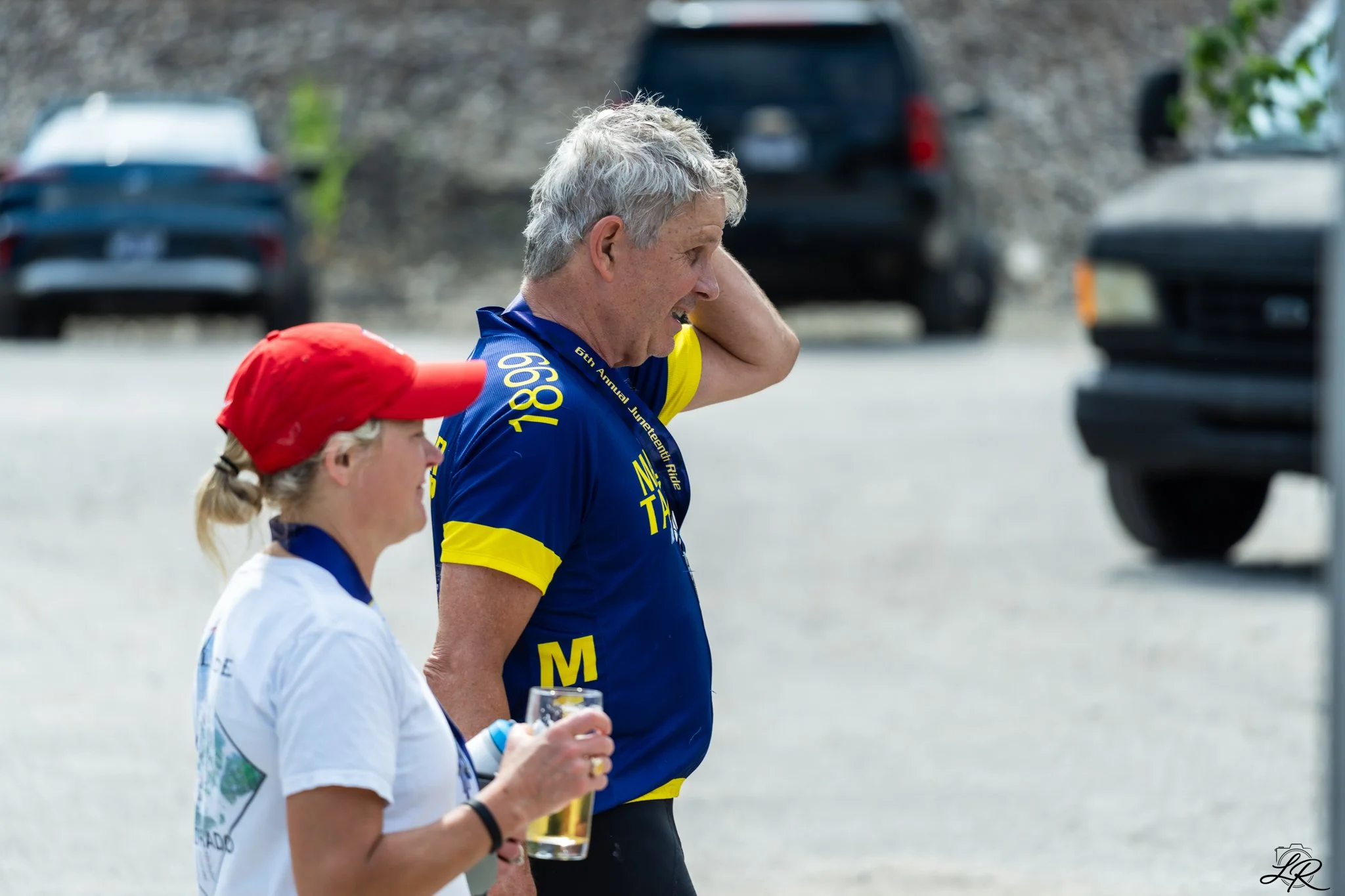 A man with gray hair in a blue and yellow shirt walking during an outdoor event, with a young girl in a white shirt and red cap walking beside him, holding a glass of drink.