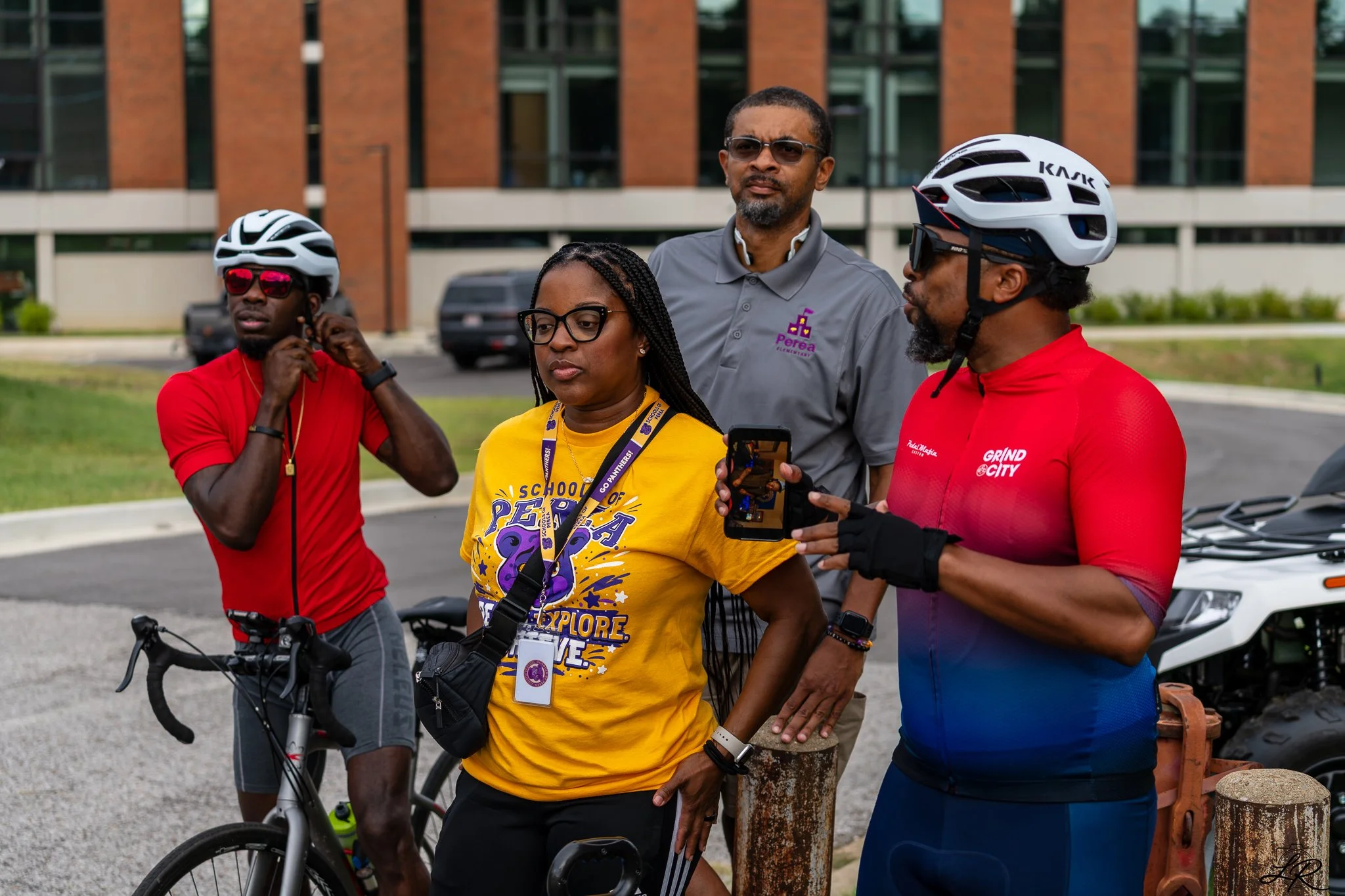 Four people standing outdoors, two wearing bicycle helmets, with bicycles and a building in the background.