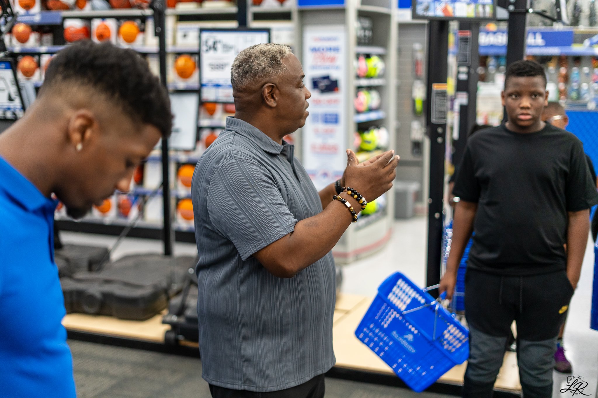 Three men at a retail store check-out counter, one is using or looking at a device, and another person is holding a shopping basket, with shelves of products in the background.