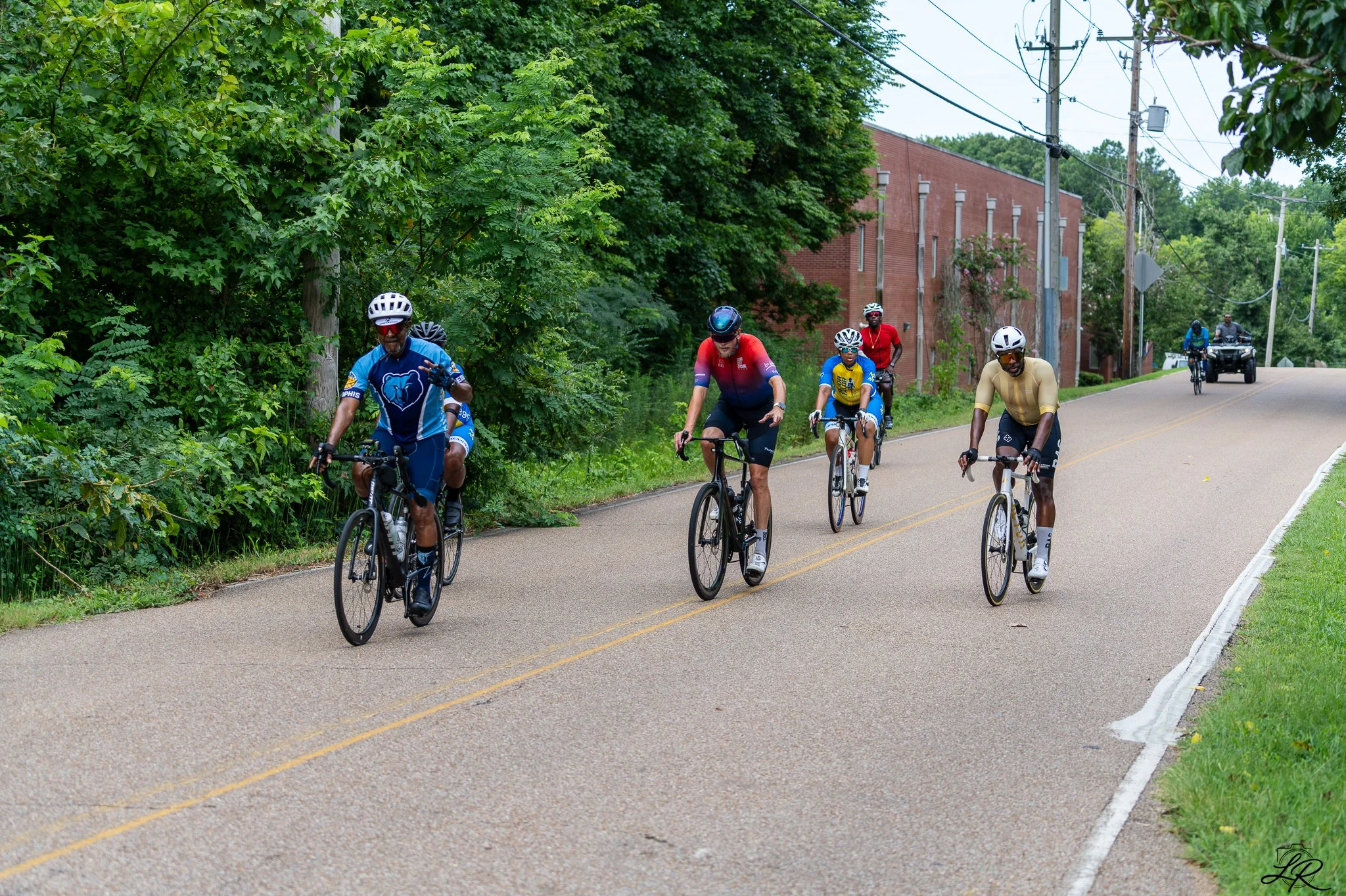 A group of six cyclists riding on a paved rural road surrounded by green trees and bushes. One cyclist is wearing a blue jersey with a bear logo, others in red, yellow, blue, and white cycling gear. Motorcycles are visible in the background, along wi