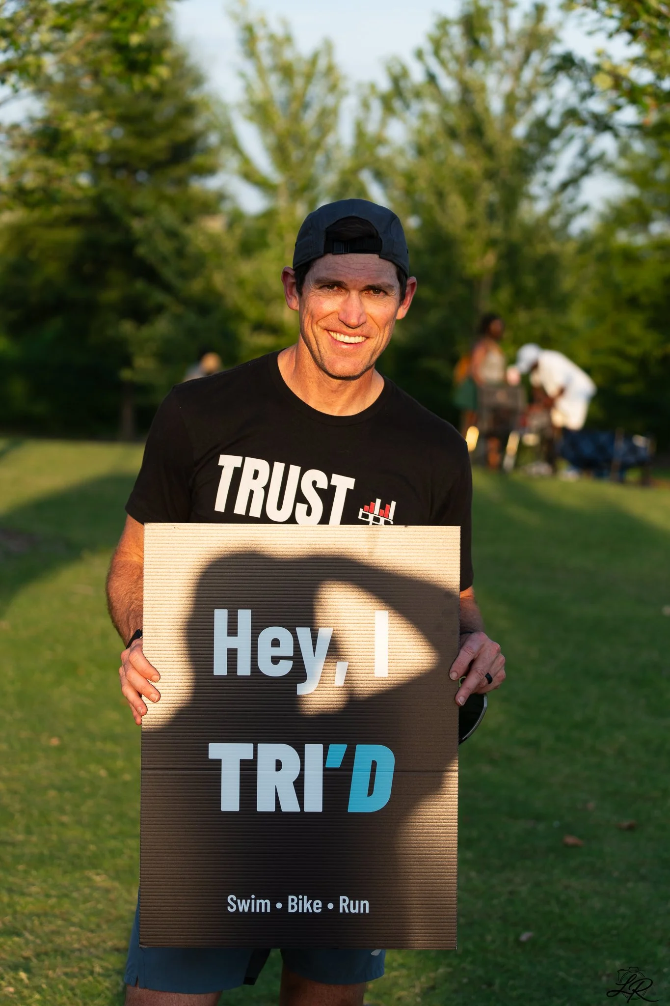 A man holding a sign that says "Hey, I TRi'D" at an outdoor event with green trees in the background, wearing a black T-shirt with "TRUST" printed on it and a black cap.
