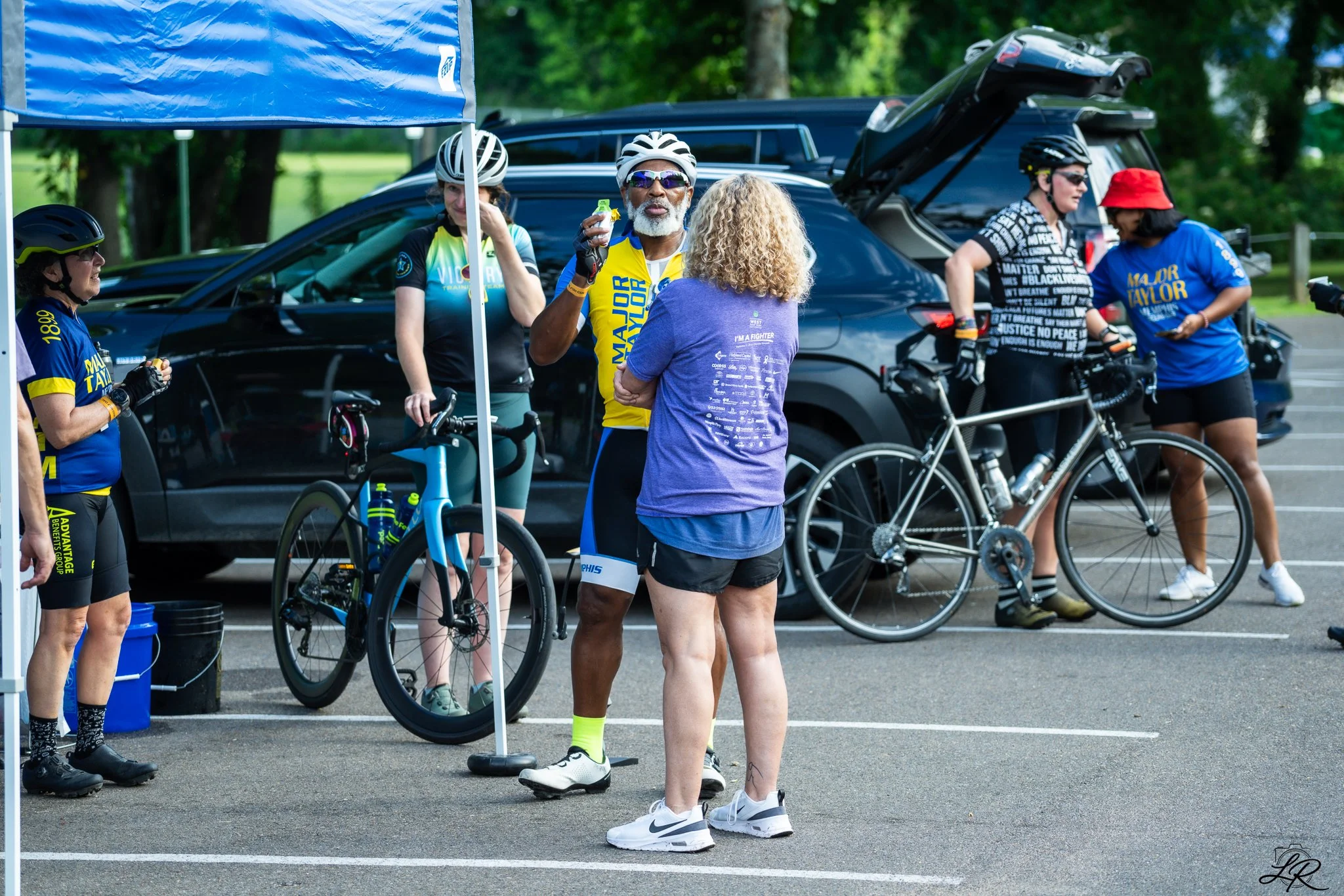 Group of cyclists and supporters gathering in a parking lot for a cycling event, with bikes and cars in the background, some people wearing helmets and colorful athletic clothing.