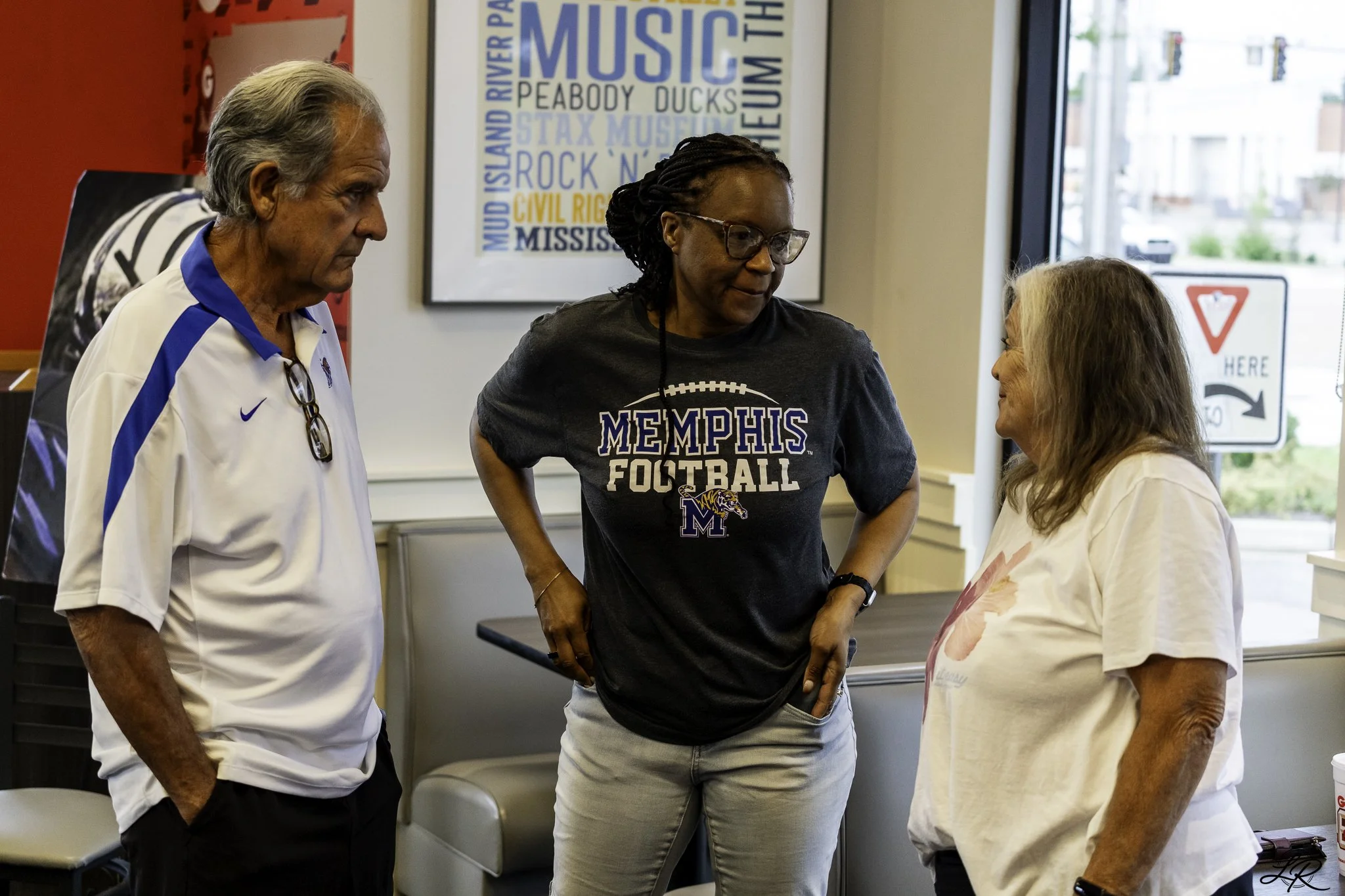 Four people are engaged in a conversation inside a room with large windows. There are two women and two men, one of the men appears to be wearing a Memphis Football t-shirt. The background features a colorful framed poster and cityscape through the w