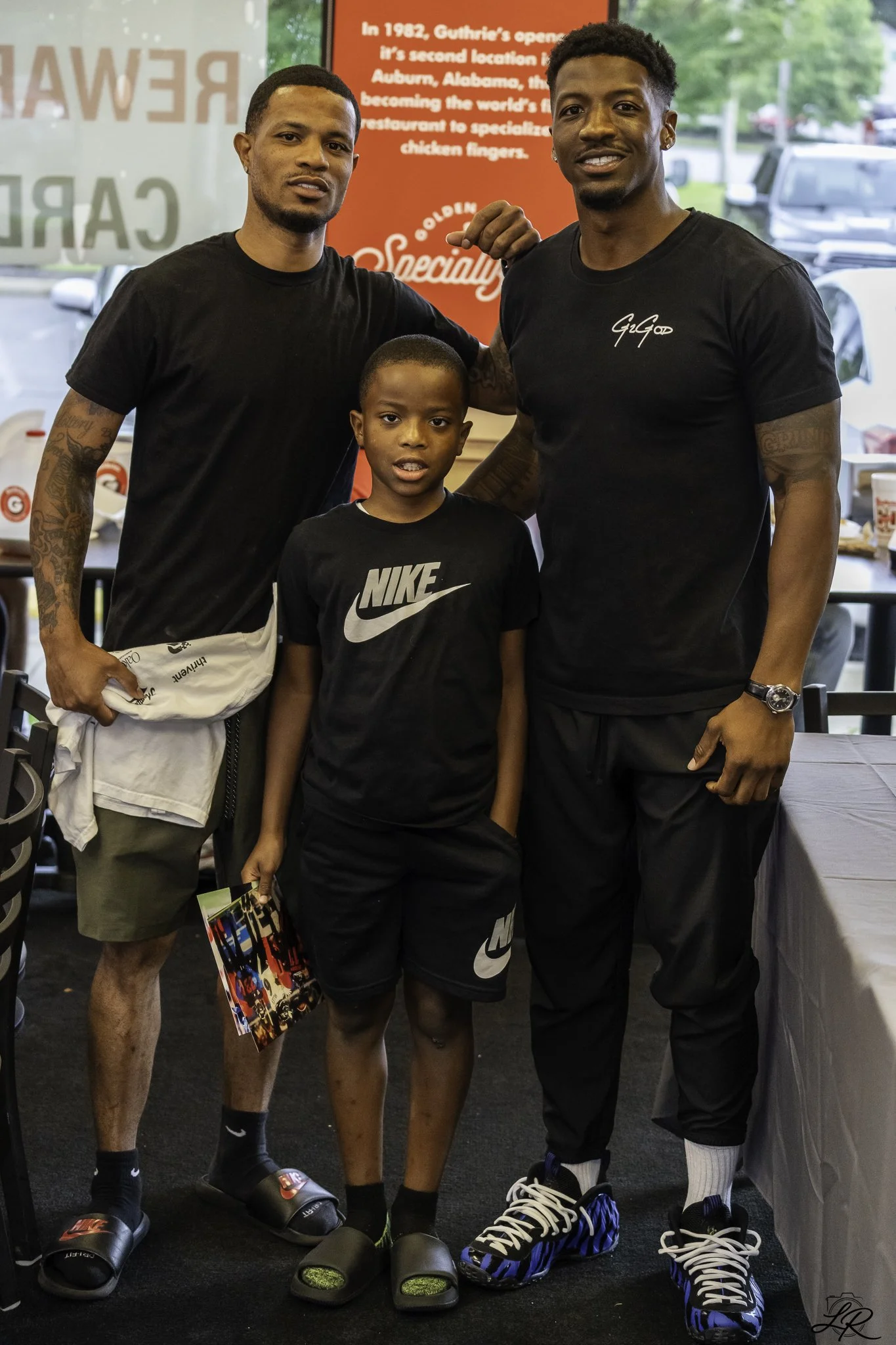 Two men and a boy standing together inside a restaurant, with a red and white sign in the background, and cars visible through the window behind them. All three are wearing black shirts and sandals or sneakers.