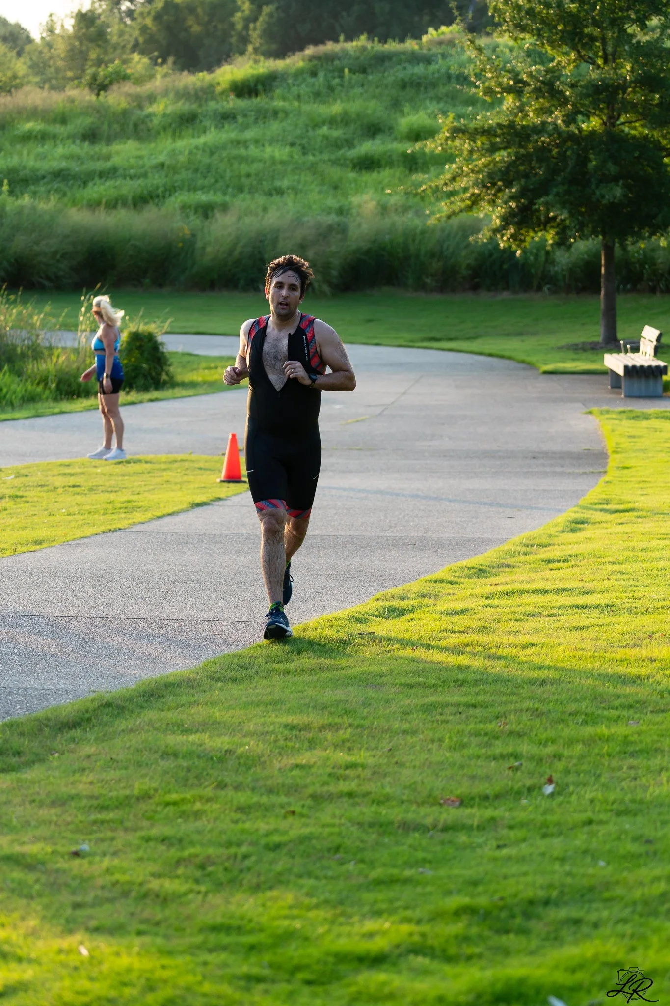 A man jogging on a paved pathway in a park with green grass and trees in the background, and a woman standing nearby.