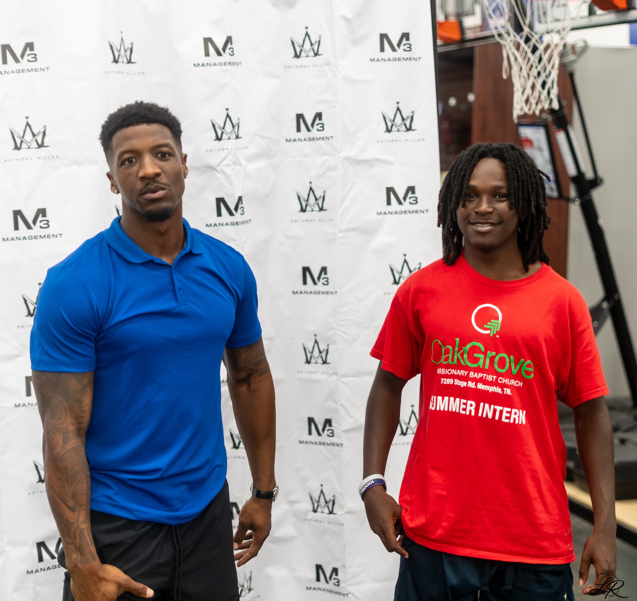 Two young men standing in front of a backdrop with logos, one in a blue shirt and the other in a red shirt, at a promotional or community event.