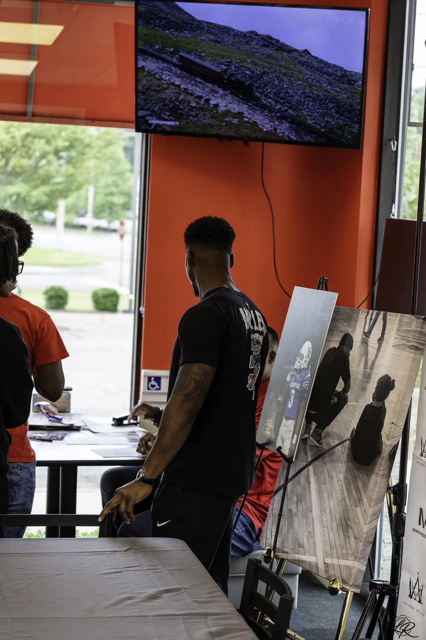 People at an indoor event with display posters, table, and TV showing landscapes, near large windows with views of trees outside.