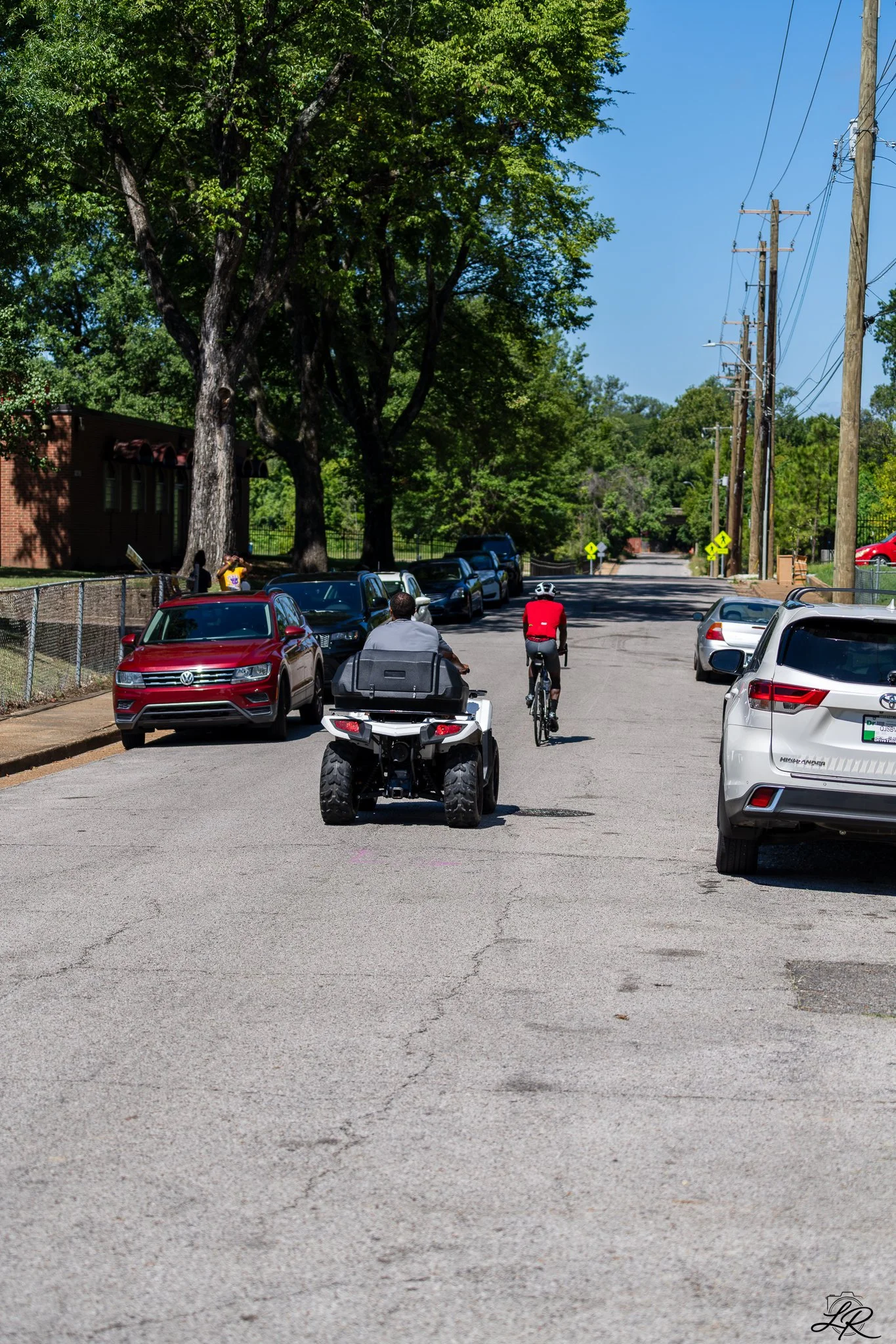 Street scene with parked cars, green trees, utility poles, and two cyclists riding down the street on a sunny day.