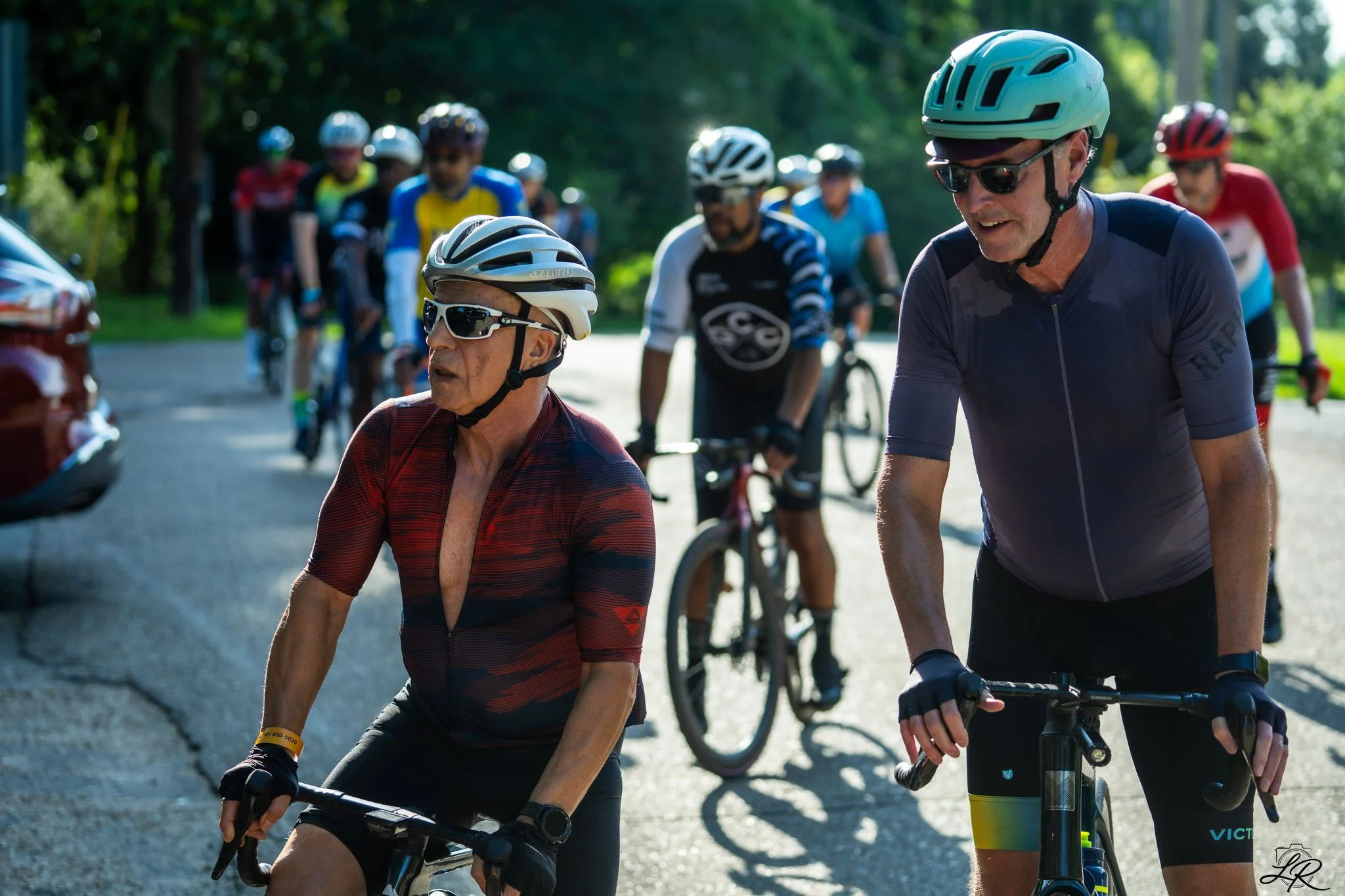 A group of cyclists, including two men at the front wearing helmets and sunglasses, riding on a shaded trail during daytime.