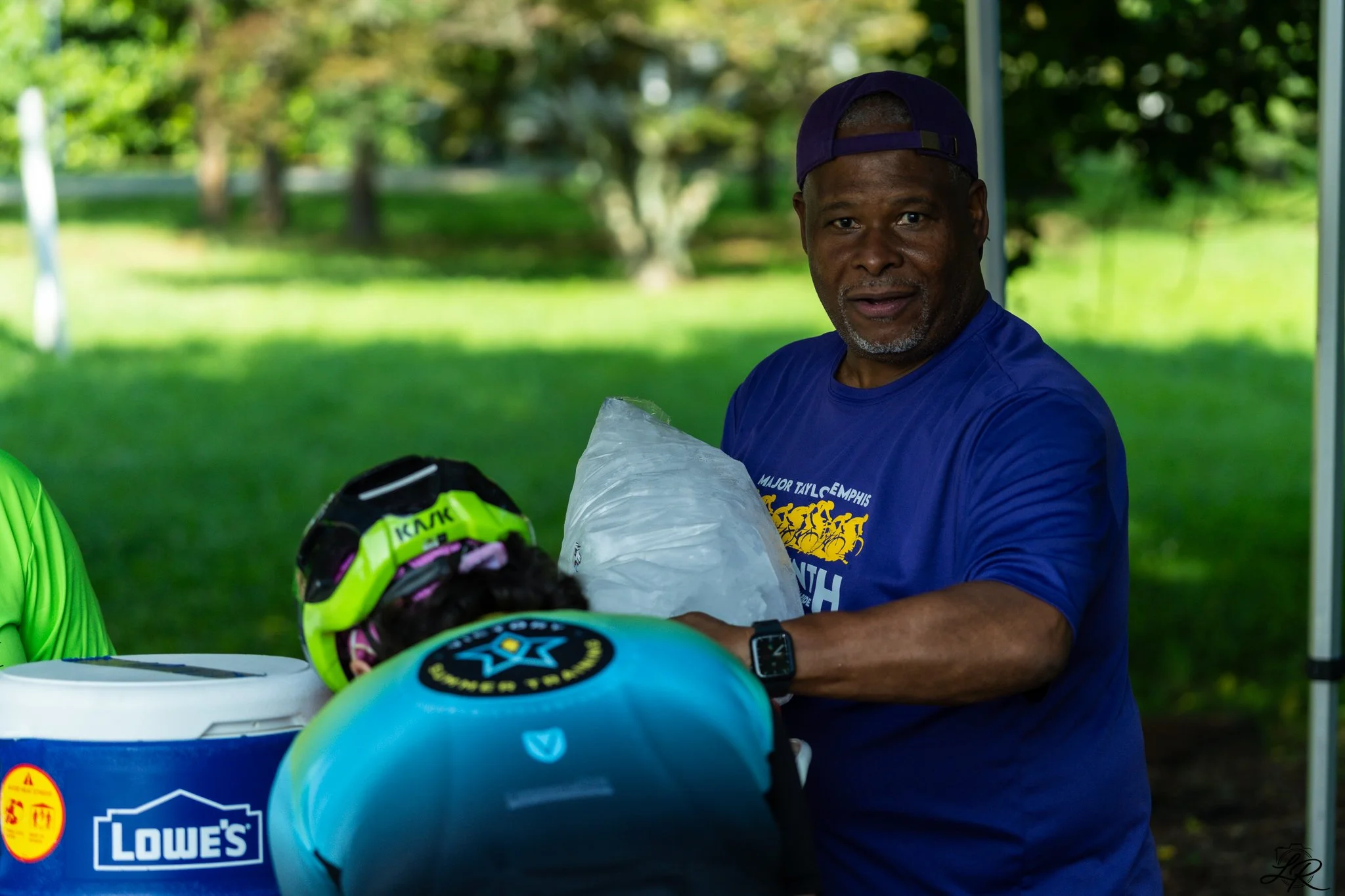 A smiling man wearing a blue shirt and a backwards cap, handling bike helmets at an outdoor event under a tent with a grassy park and trees in the background.
