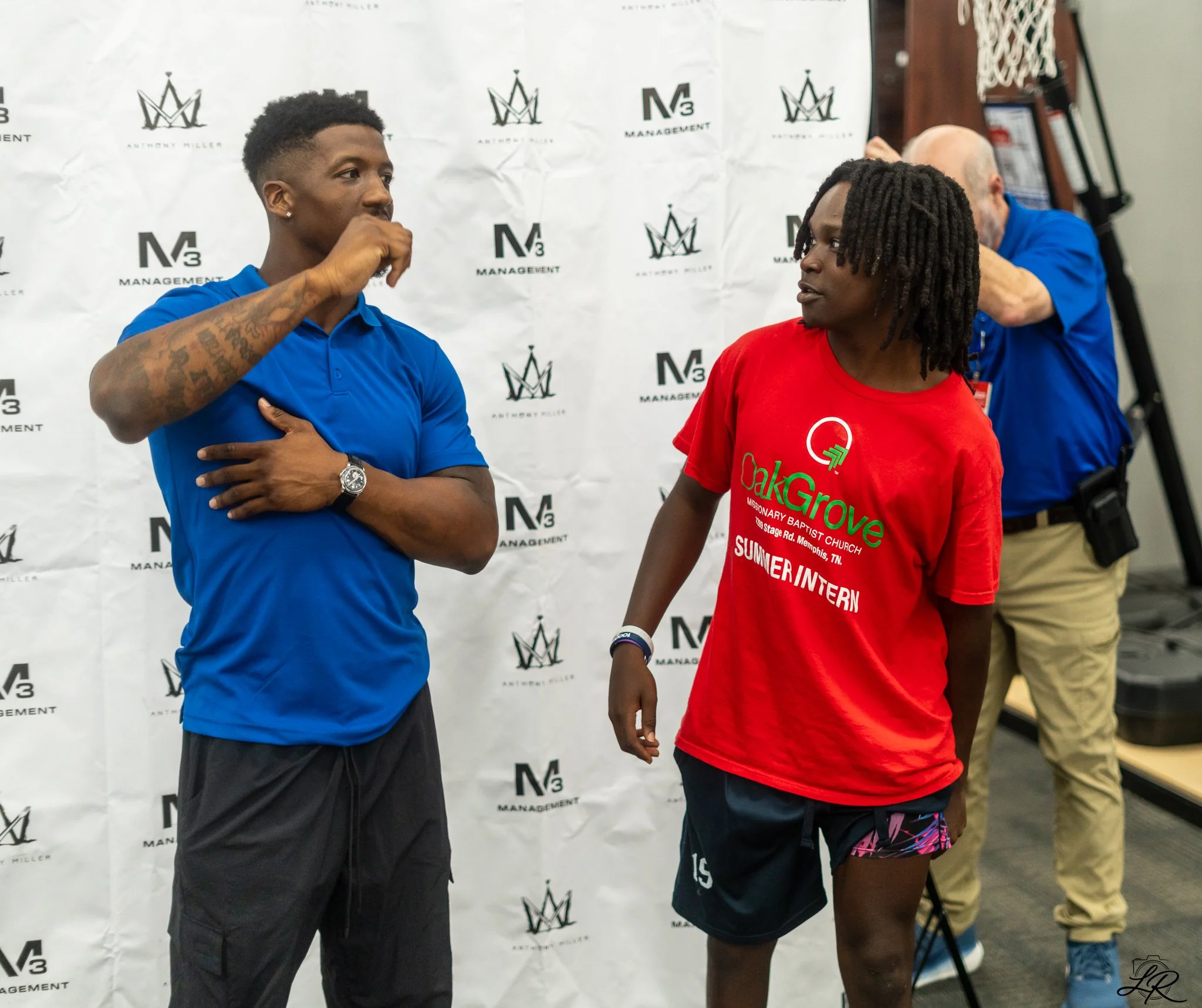Two young men are standing and facing each other in front of a white backdrop with logos and text, one wearing a blue shirt with tattoos and the other wearing a red t-shirt with event info. The man in blue has his hand on his chin and crossed arms, w