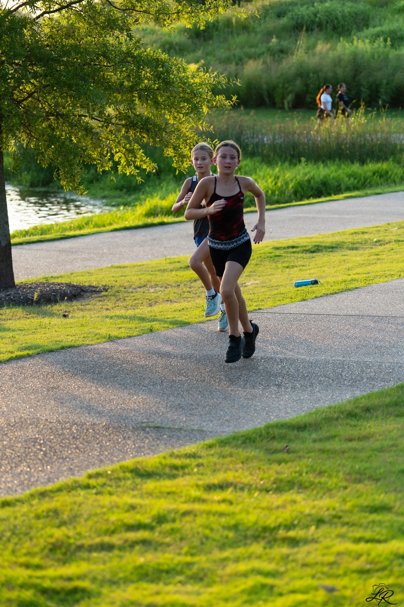 Two young girls running along a paved path near a green grassy area and a small waterway, with trees and a creek in the background during the daytime.