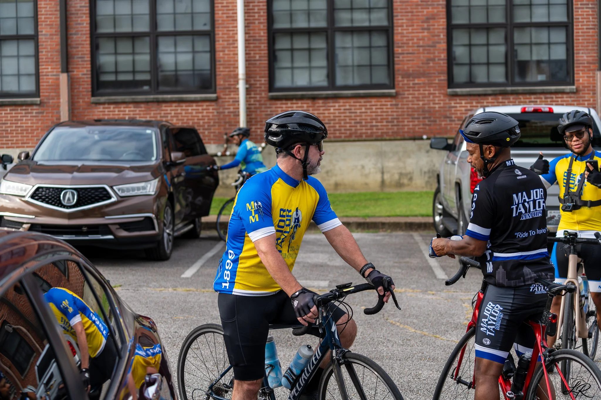 Group of cyclists wearing blue and yellow jerseys and black helmets, standing with bikes in a parking lot with cars and a brick building in the background.