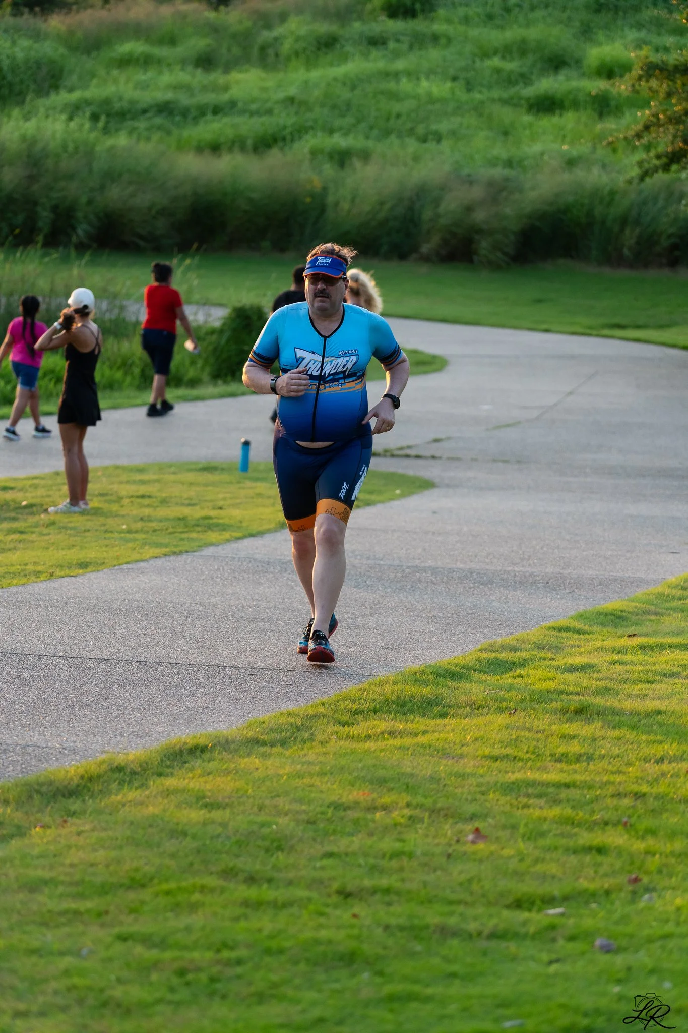 A man running on a park trail, wearing a blue sports shirt and shorts, a visor, and sunglasses, with a group of people walking in the background, greenery and trees surrounding the area during daytime.