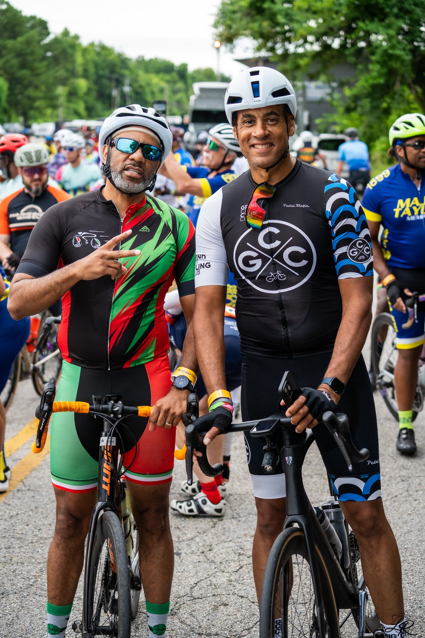 Two men with bicycles smiling at the camera at a cycling event, surrounded by other cyclists in colorful gear, outdoors on a cloudy day.