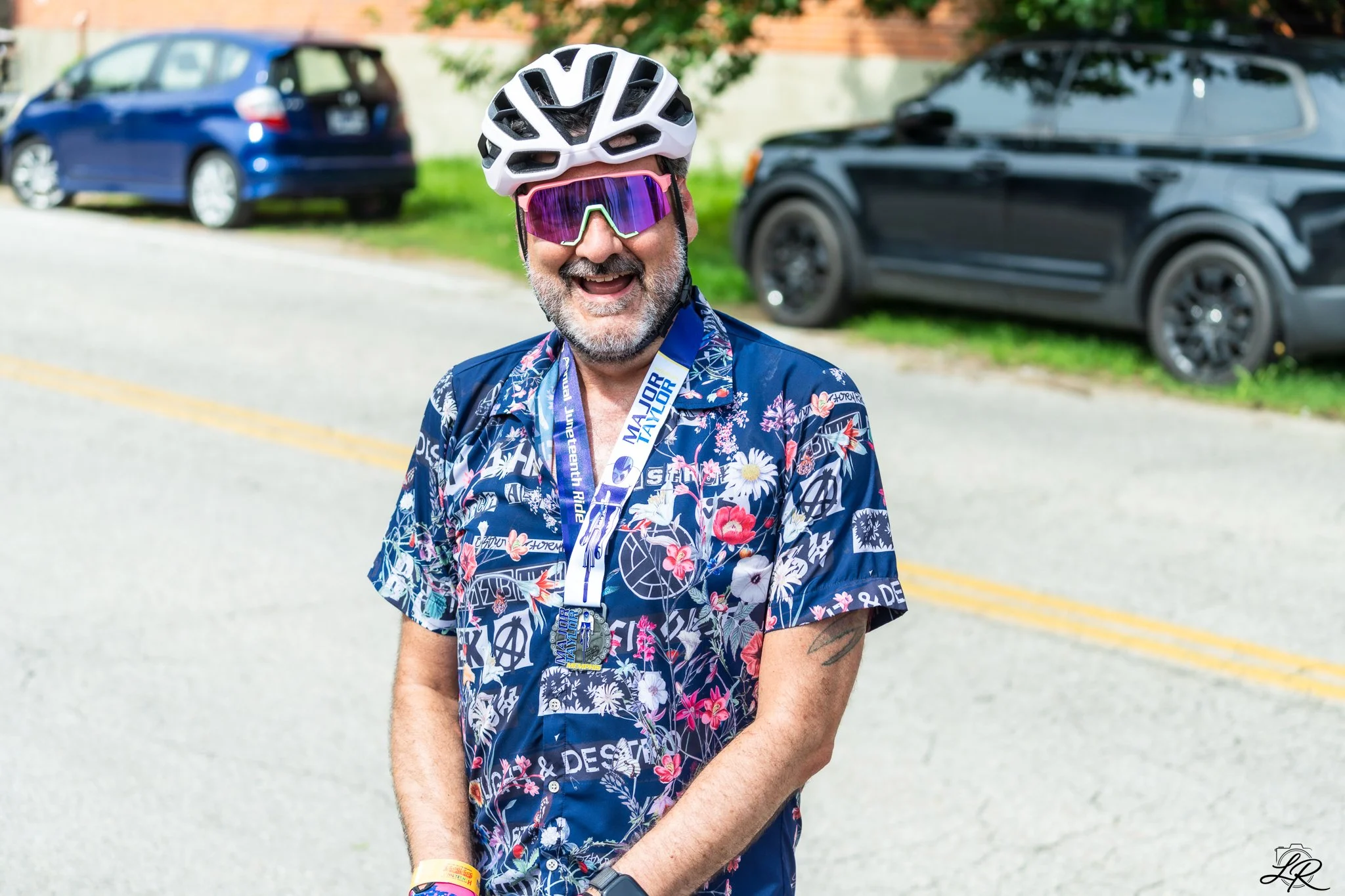 A smiling man in a floral Hawaiian shirt wearing a white bike helmet and purple sports sunglasses outdoors, with cars and greenery in the background.