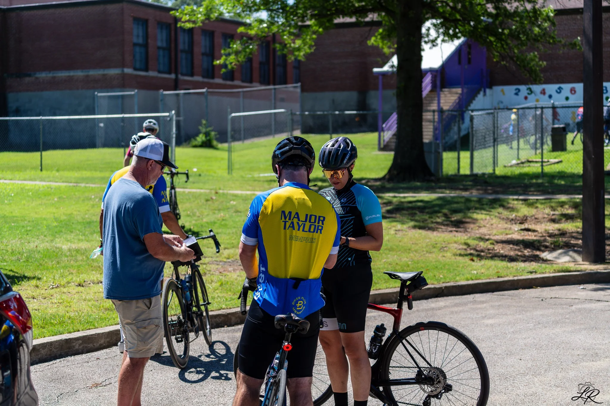Group of cyclists and a man with a notepad outside on a sunny day, with a playground and a brick building in the background.