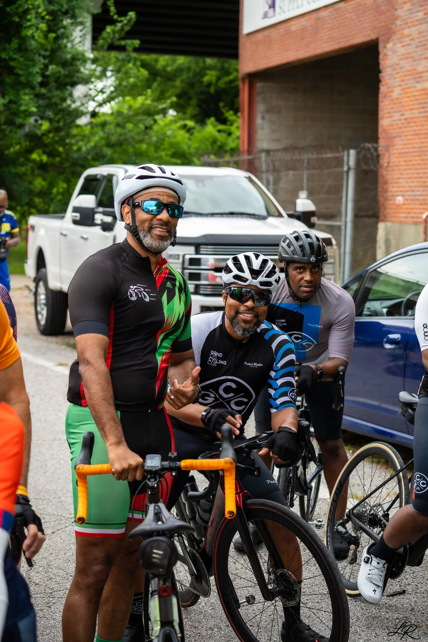 Group of cyclists with helmets and sunglasses, standing with their bikes outside near a white truck and brick building.