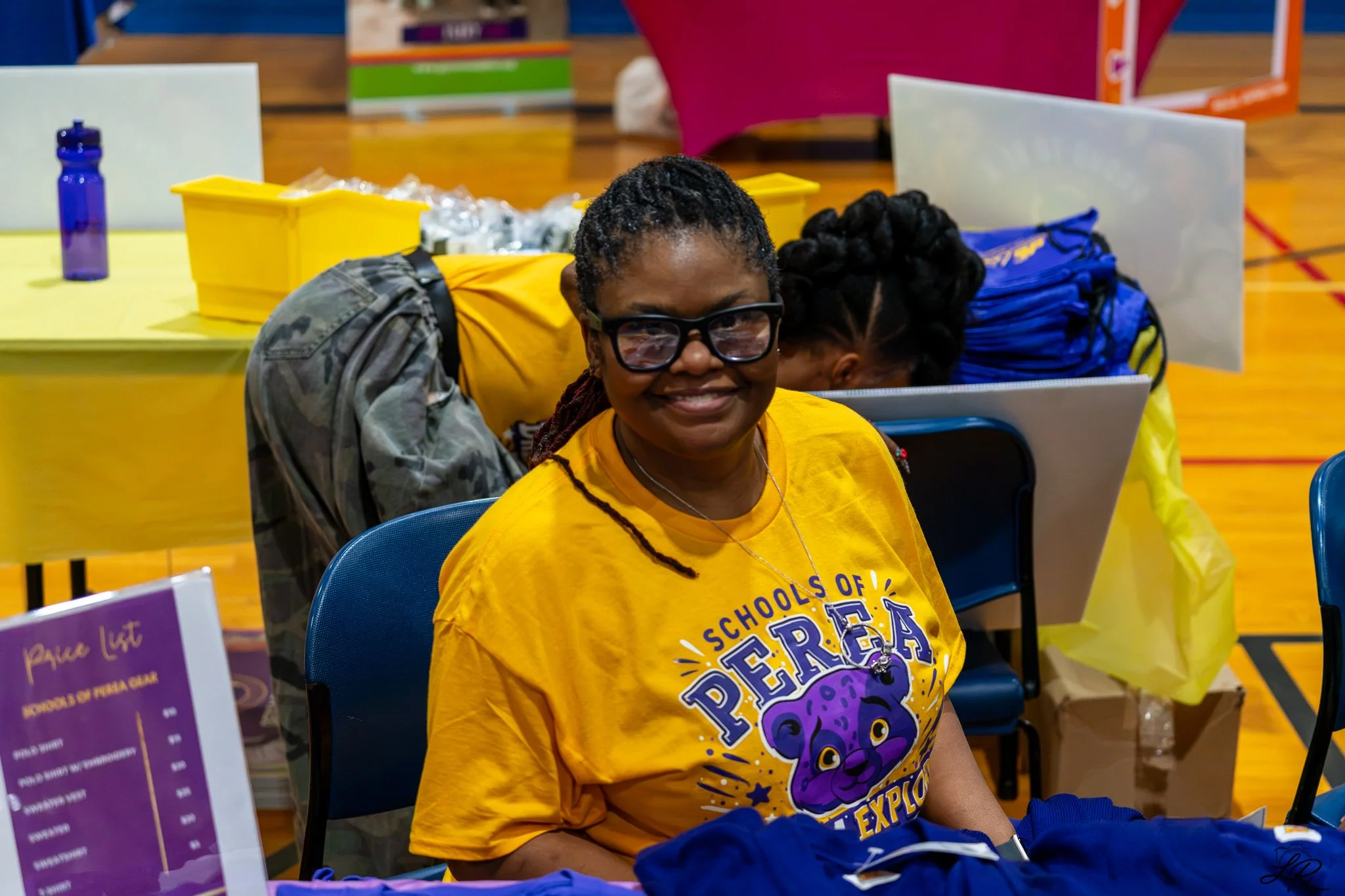 A woman with braids, glasses, and a bright smile, wearing a yellow T-shirt with a purple and yellow design, sitting at a table during an event in a gymnasium. Behind her, there are tables with various items and other people, and the event appears che