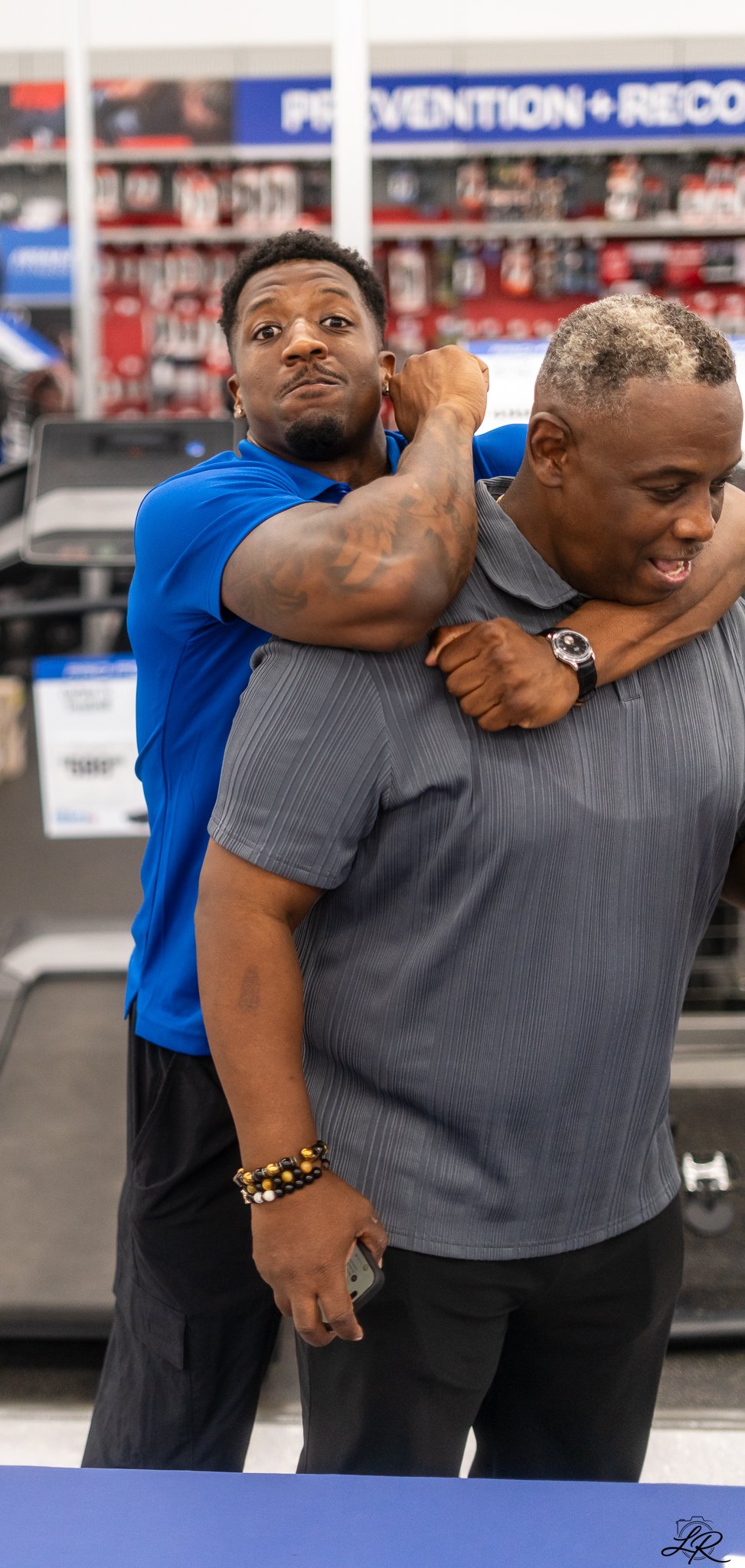 A man with short, gray hair and a goatee wearing a gray shirt, carrying a younger man with short black hair and a beard, wearing a blue shirt, on his back in a store with a sign that reads "PRESENTATION + RECORD."