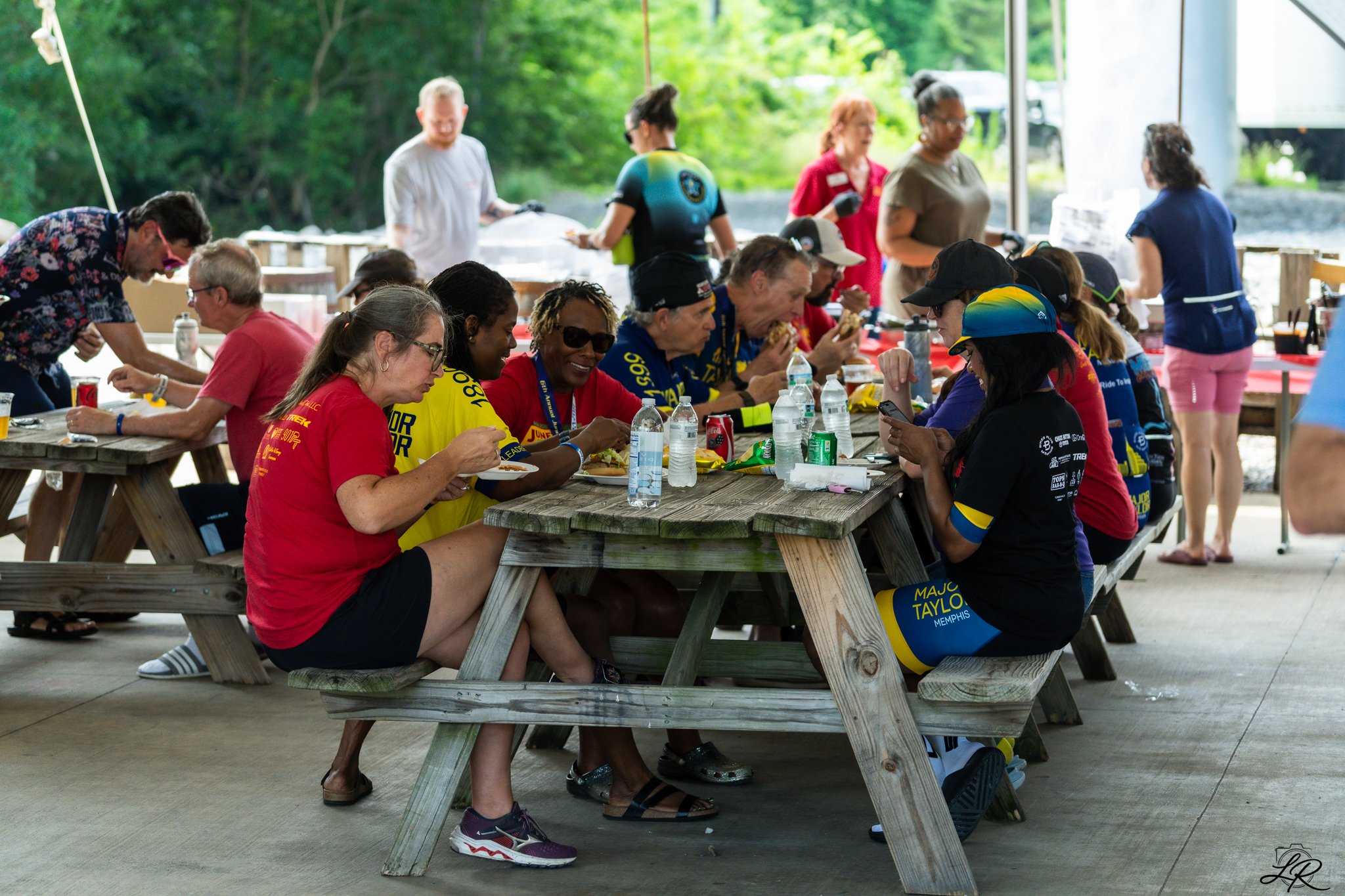A group of people sitting at a picnic table eating and drinking, with more people standing and talking in the background under a pavilion with a view of trees.