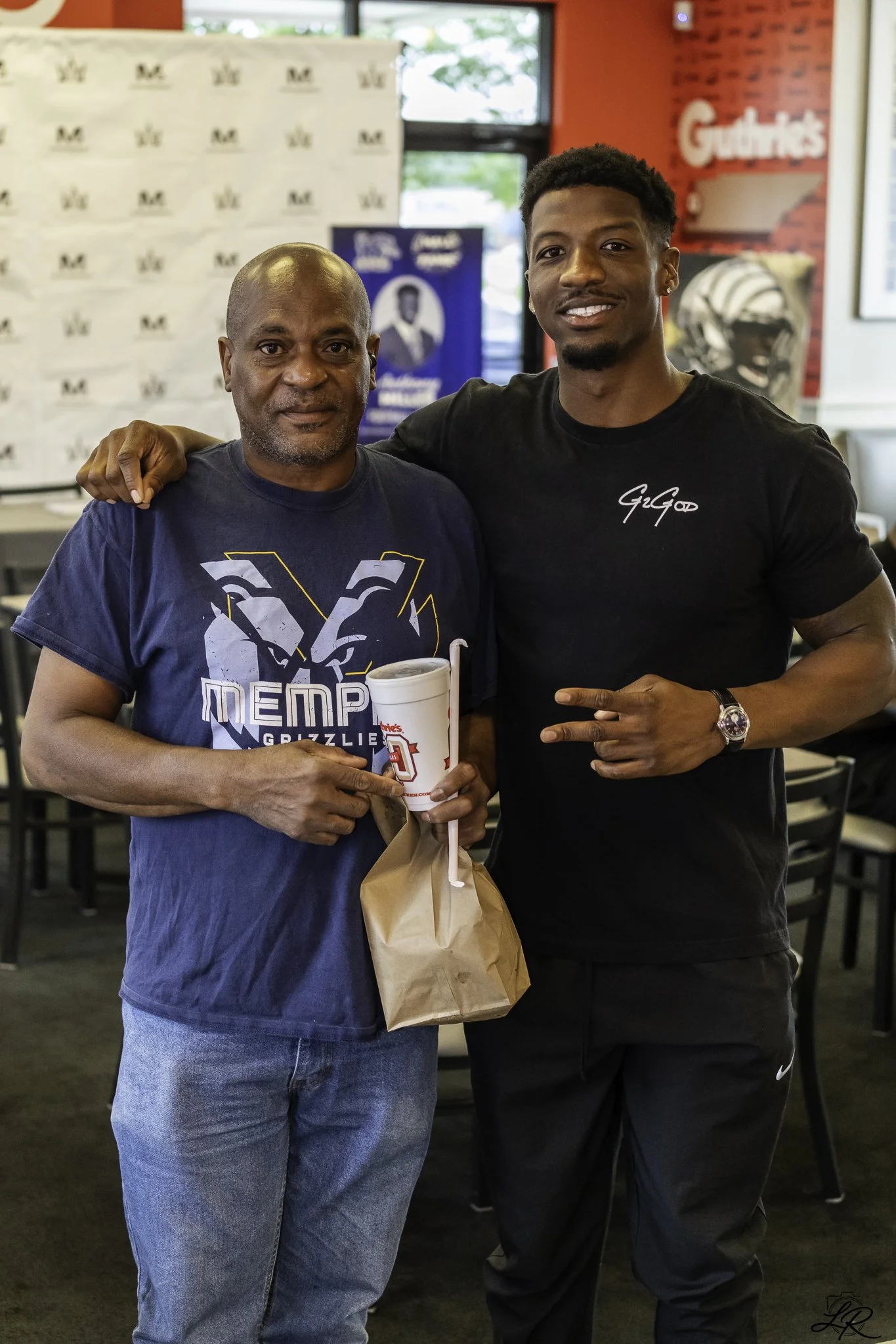 Two men standing in a restaurant, one older with a shaved head holding a drink, and the younger man with short hair dressed in a black t-shirt, smiling for the photo.