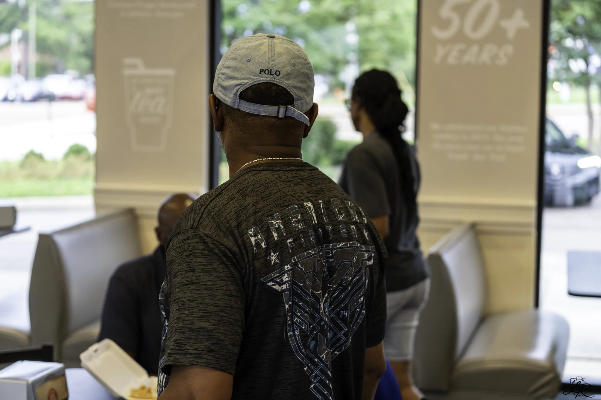 A man wearing a light grey baseball cap with the word 'POLO' on it, and a dark t-shirt with a graphic design, standing indoors with his back to the camera. In the background, there is a woman with dark hair and glasses, wearing a dark jacket, standin