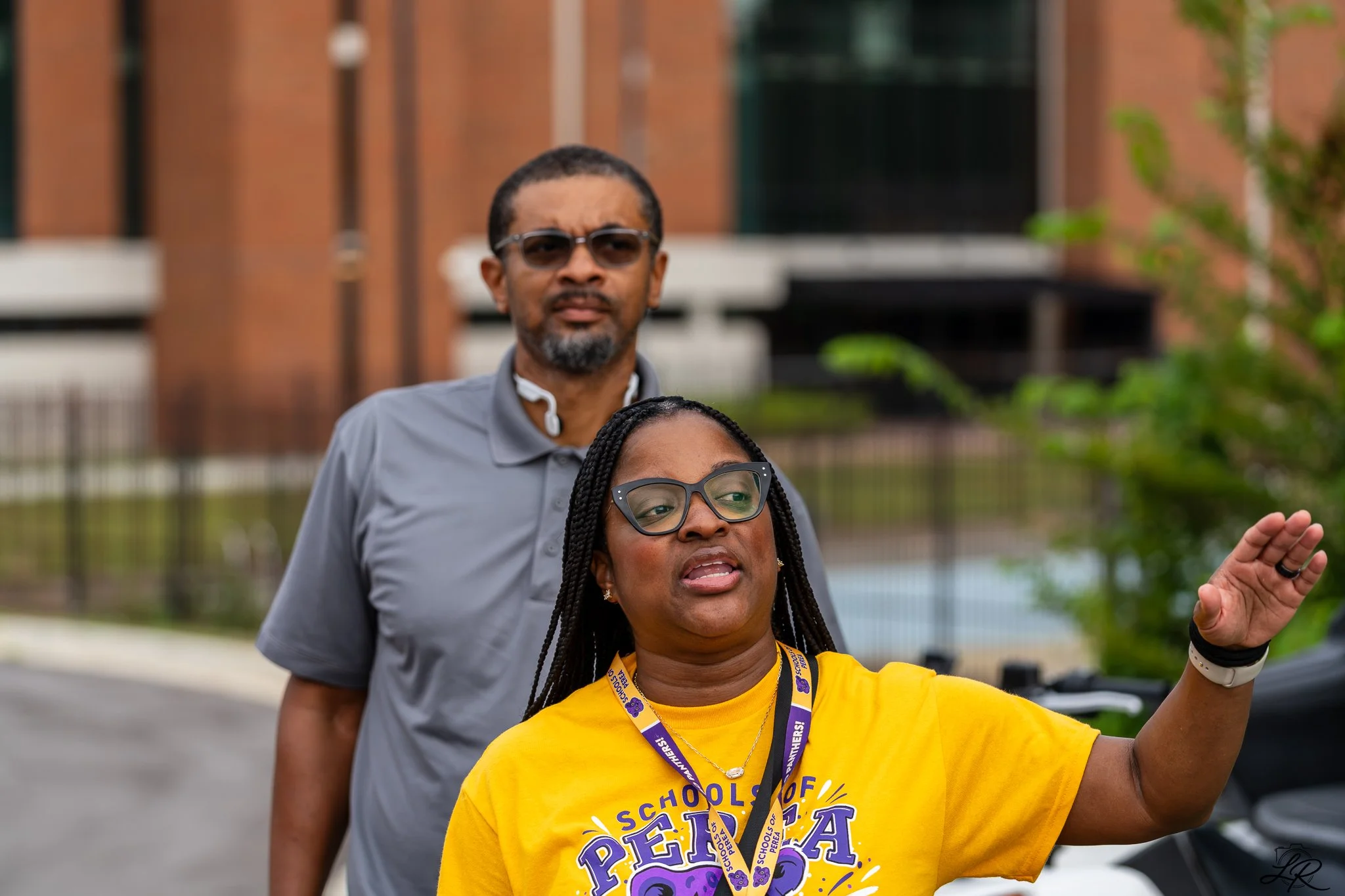 Two women and a man outdoors with a brick building in the background. The woman in the foreground is wearing glasses, a yellow shirt, and a purple lanyard. The man behind her is wearing sunglasses and a gray shirt. The woman in the foreground is spea
