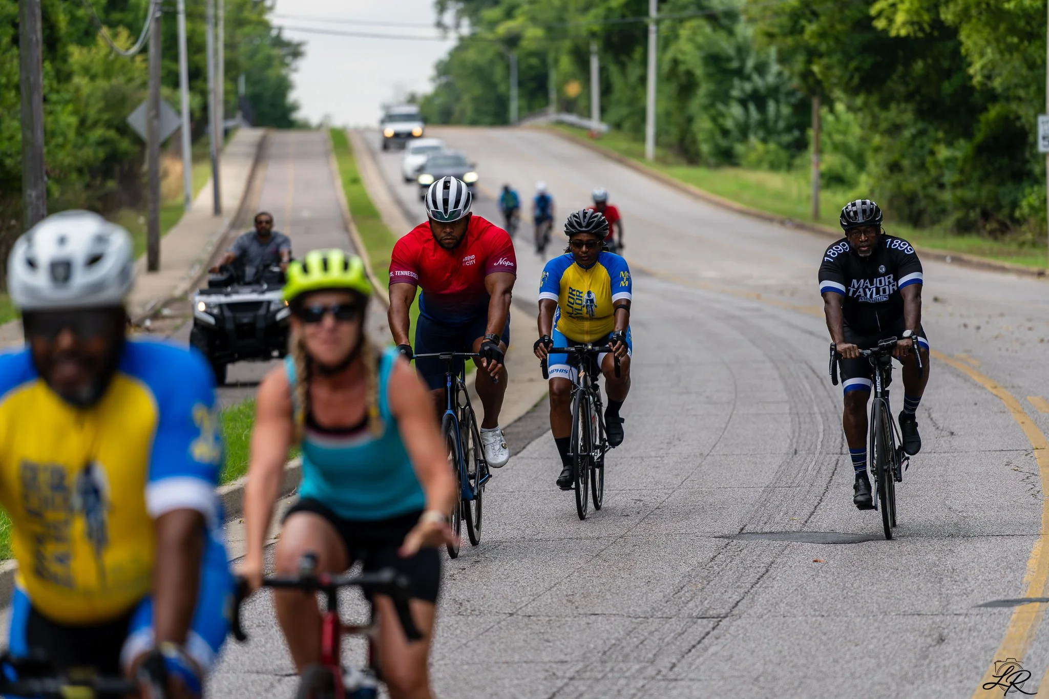 A group of people, mostly wearing helmets, riding bikes on a road with cars and greenery in the background.