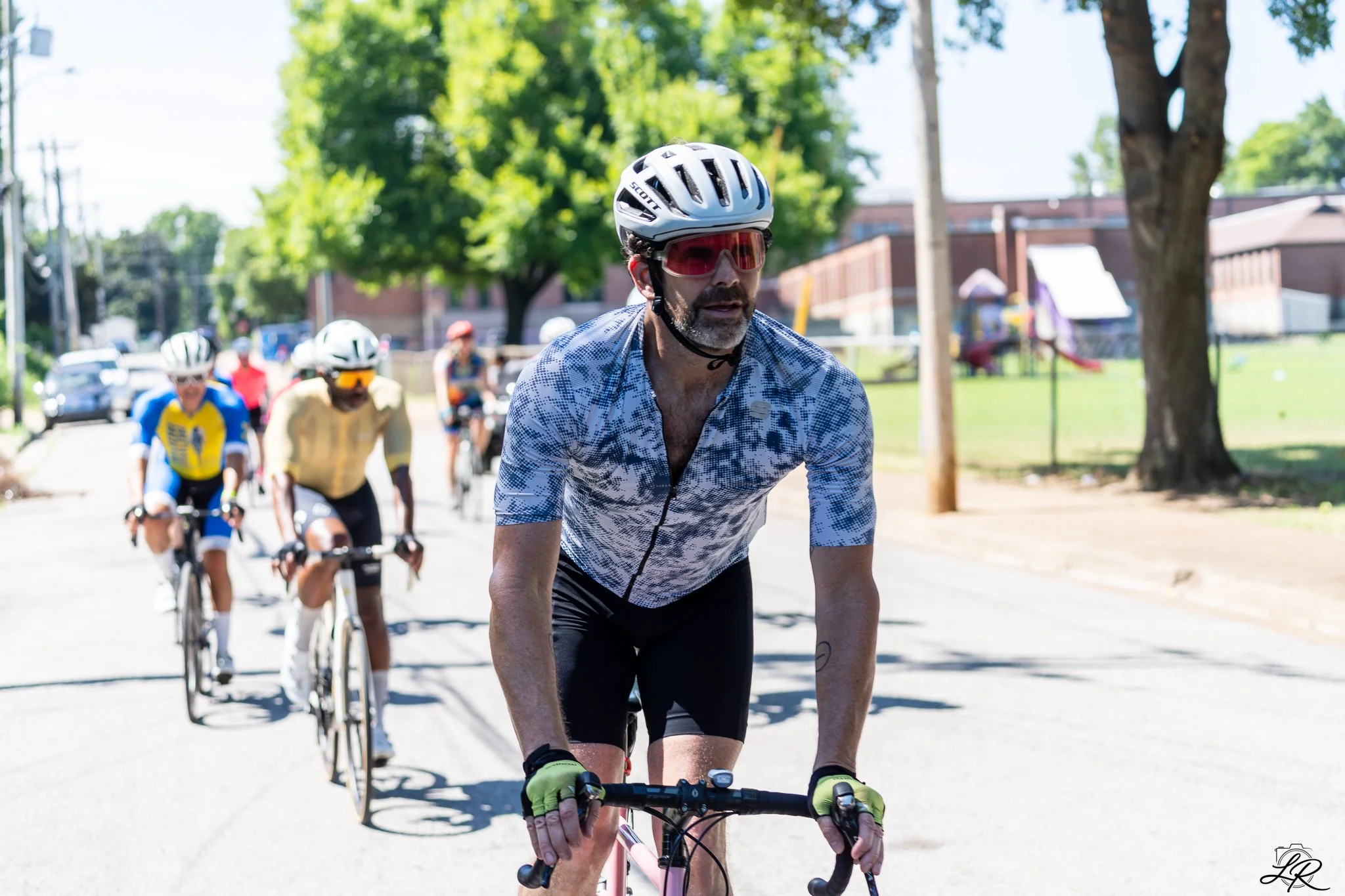 A group of cyclists riding on a sunny day, with a man in the front wearing a helmet and sunglasses, dressed in a patterned cycling jersey, surrounded by trees and spectators in the background.