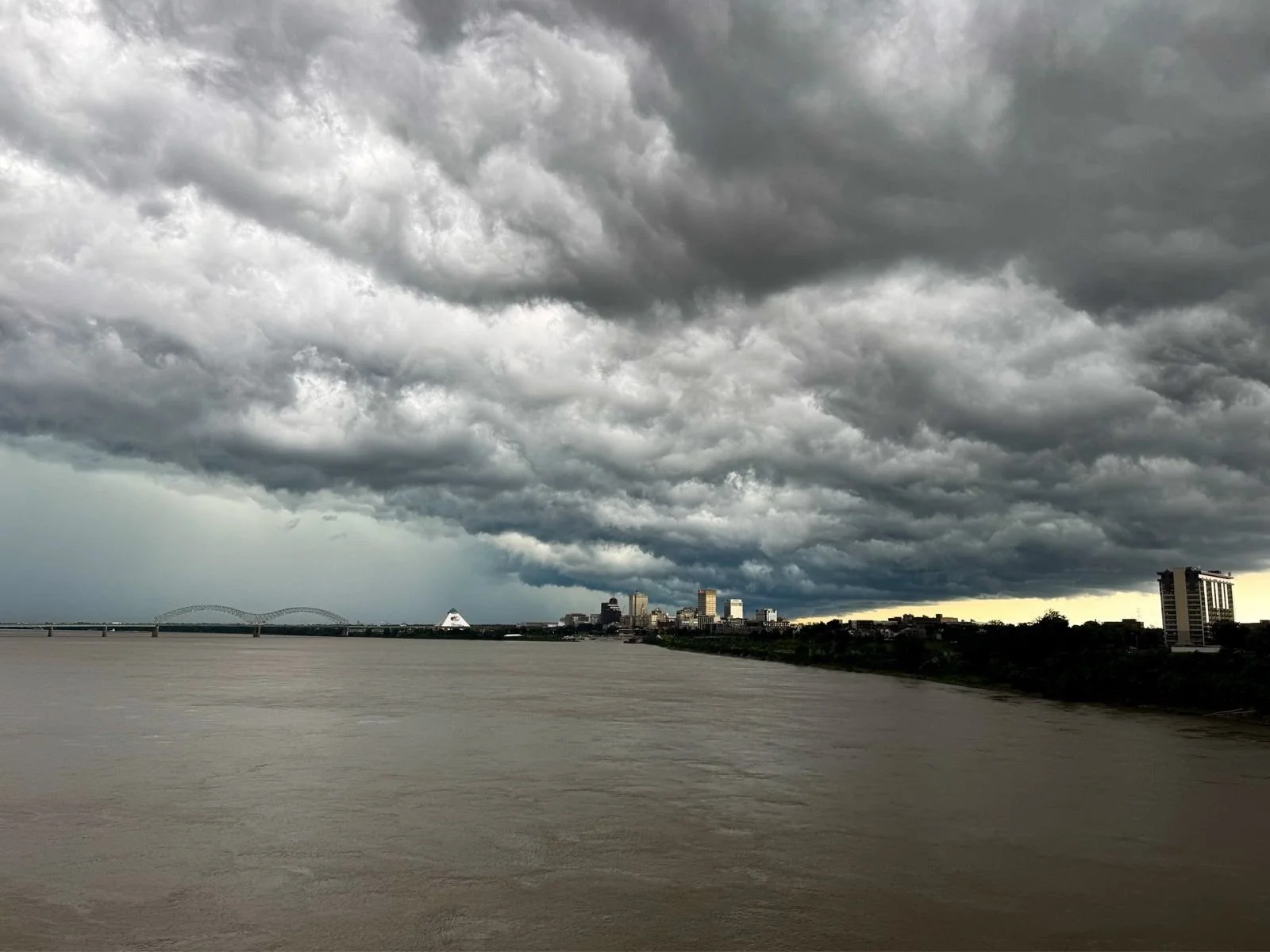 Dark, stormy clouds over a river with a city skyline in the distance, including a bridge and a pyramid-shaped building.