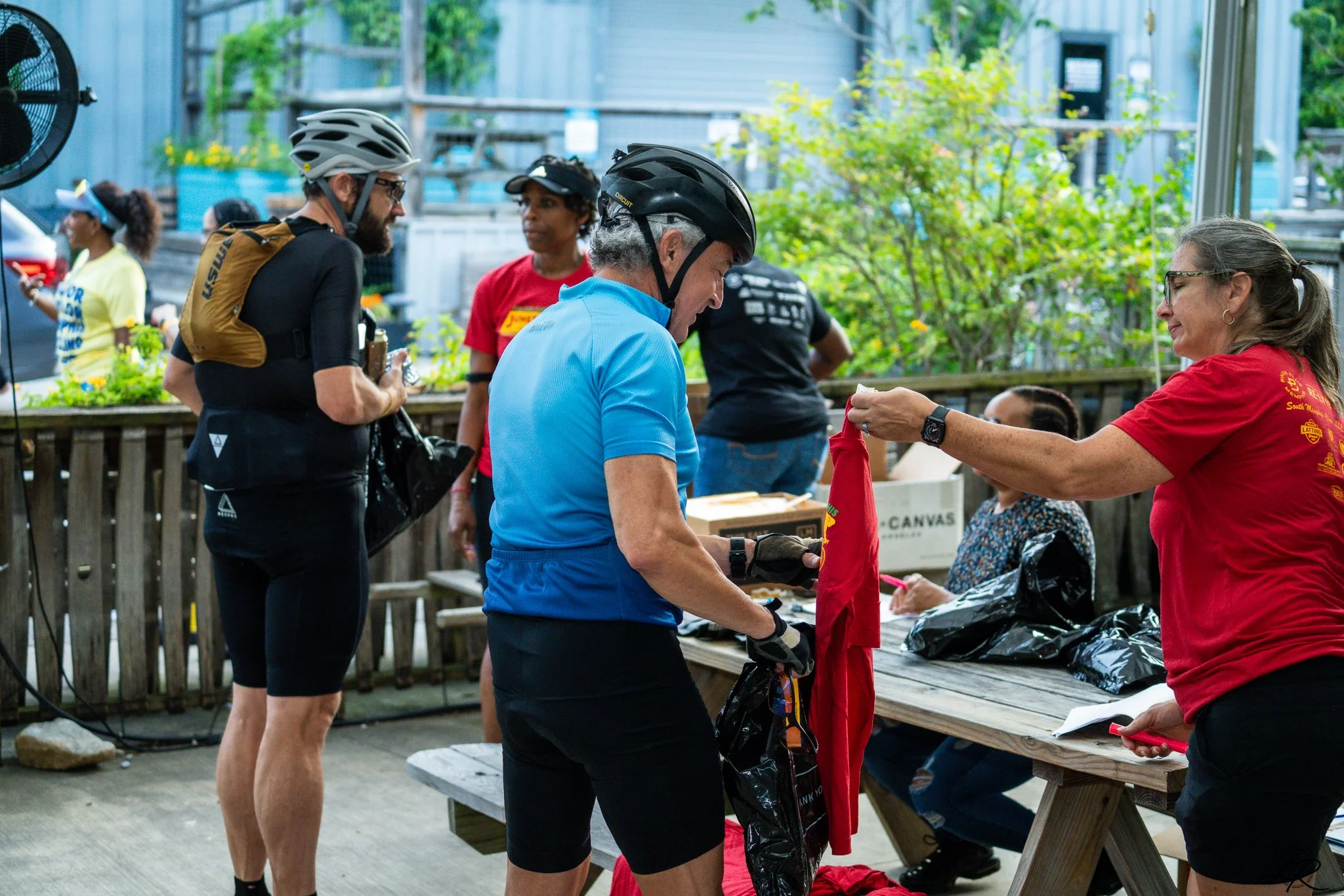 People at an outdoor event, with some wearing bicycle helmets peeling off and others holding items behind a table. The setting appears to be a gathering or celebration with a green background and wooden fencing.