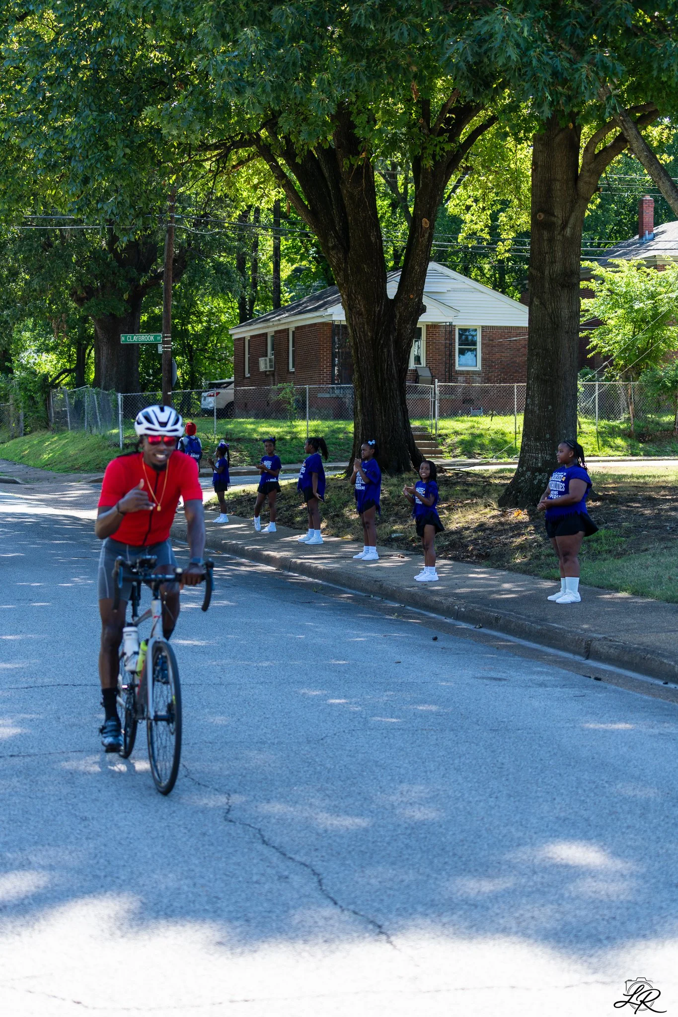 A man in a red shirt and helmet riding a bicycle on a residential street while seven girls in blue shirts and black shorts watch from the sidewalk next to tall trees and a house.