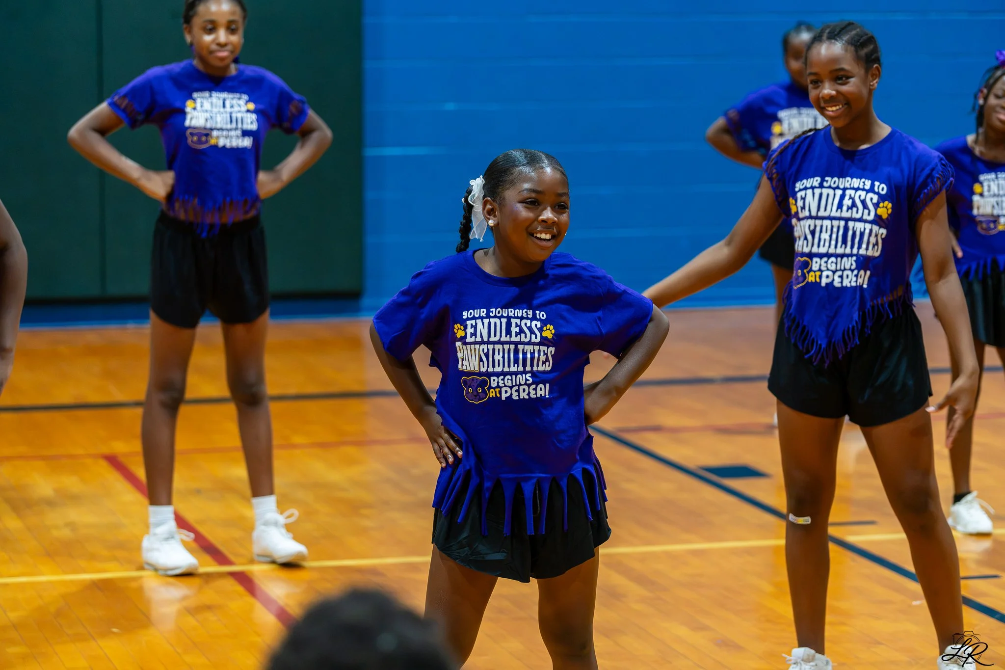 Group of young girls in blue shirts and black skirts standing on a gymnasium floor, smiling and posing with their hands on hips, participating in a dance or cheer practice.