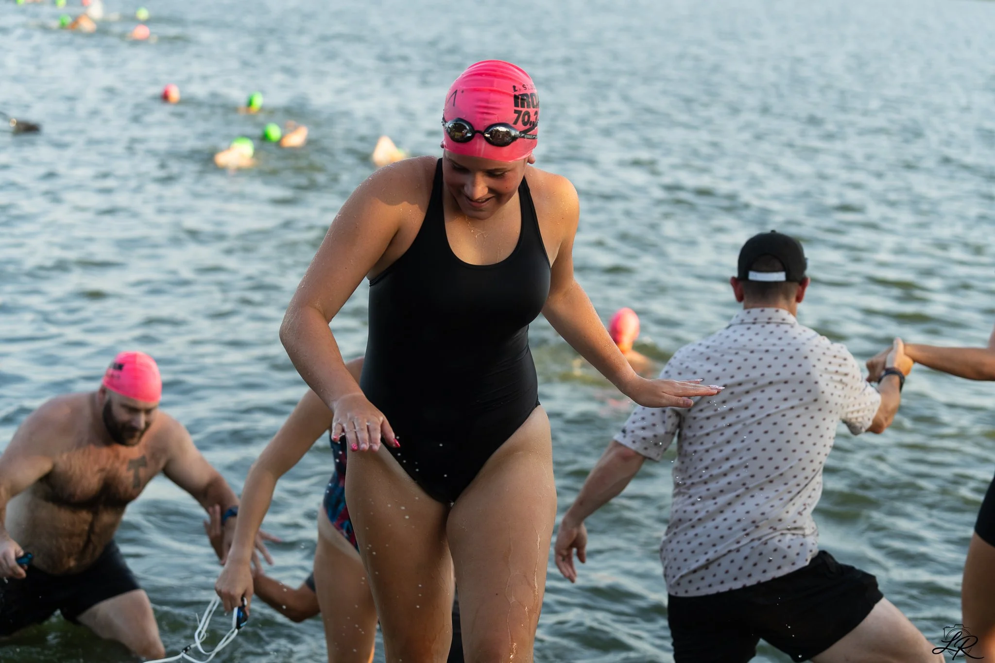 A group of swimmers, including a woman in a black swimsuit and pink swim cap, are entering a body of water, possibly for a race or event, with buoys floating in the water.