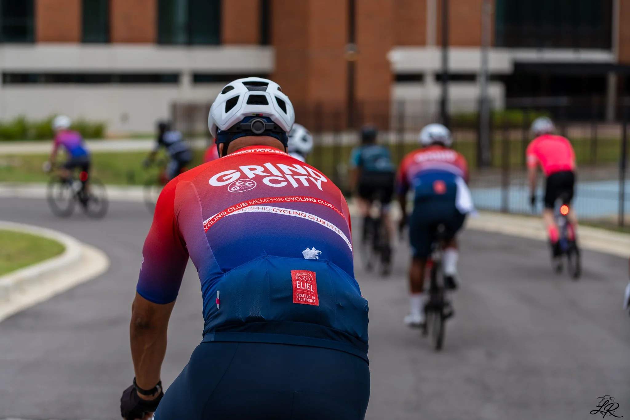 Back view of a cyclist in a red and blue jersey riding in a group of cyclists on a paved road near a building with a brick exterior and large windows.