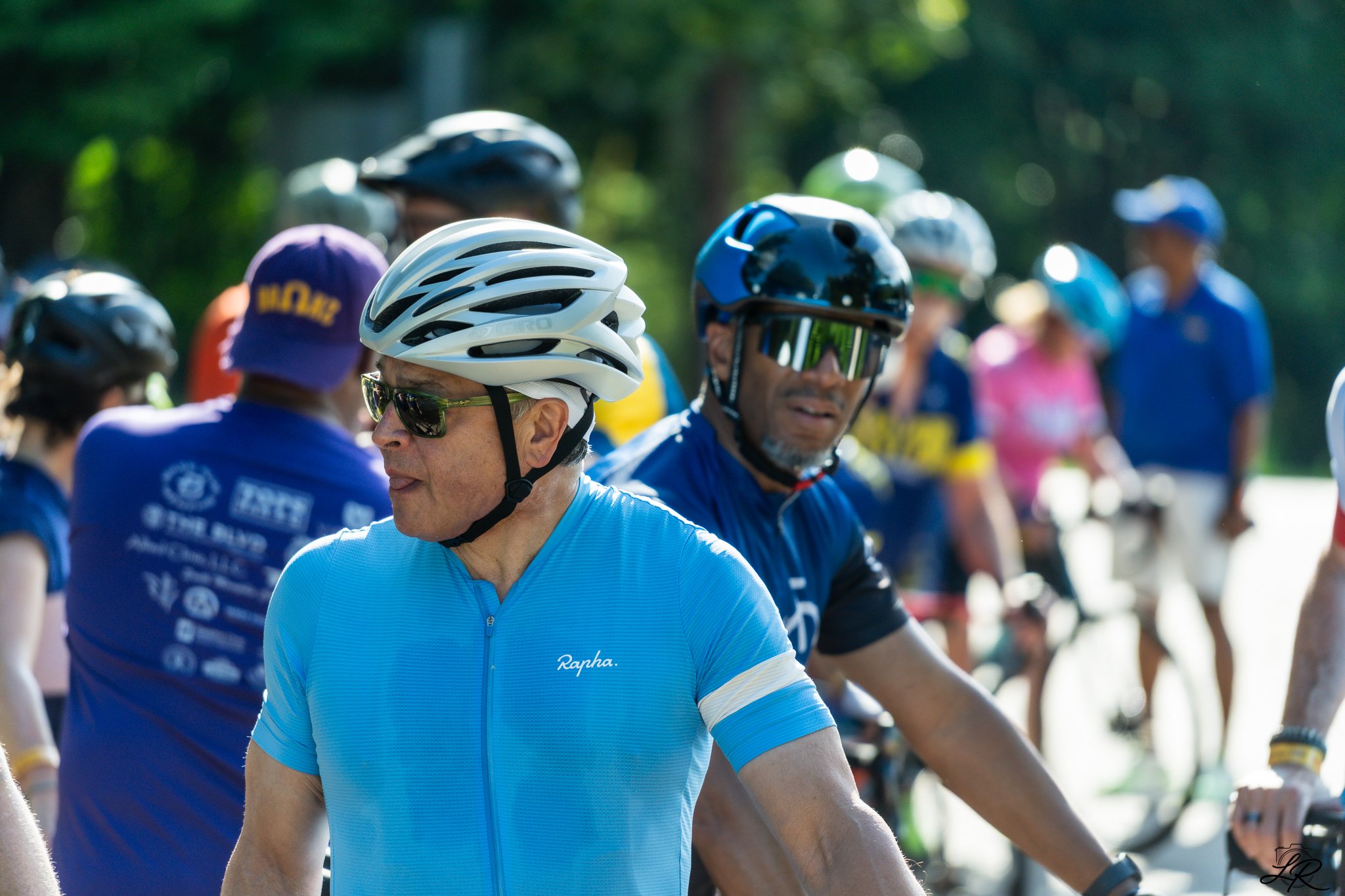 Group of cyclists wearing helmets and sunglasses during a race or event, with some in blue jerseys and others in various colors, outdoors with greenery in the background.
