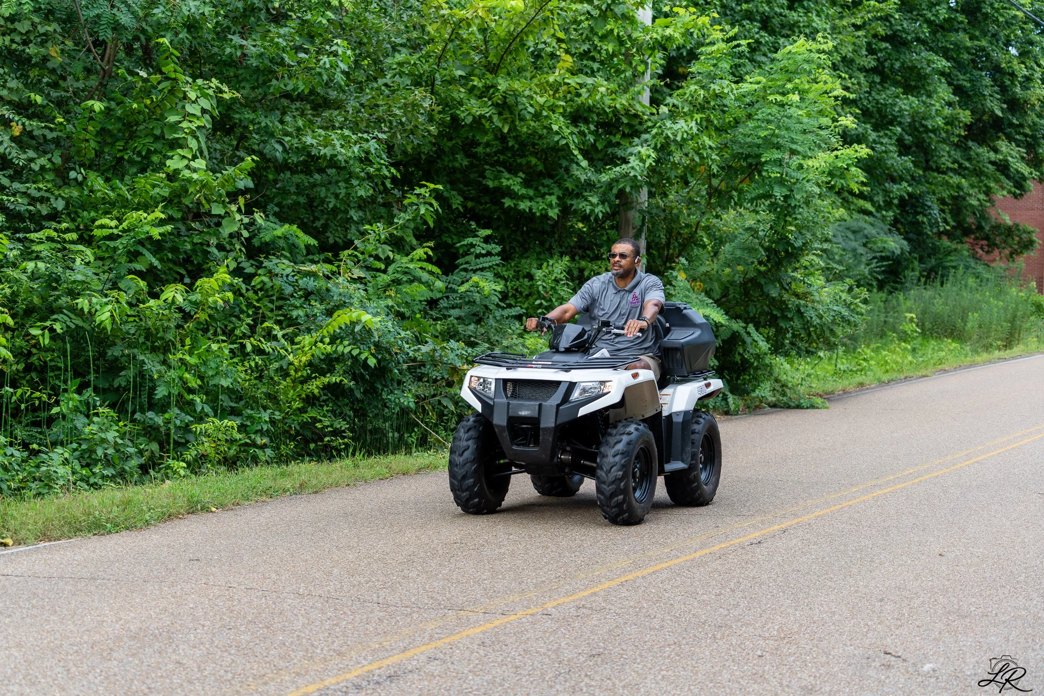 A man riding a white all-terrain vehicle on a paved road, with lush green trees and bushes in the background.