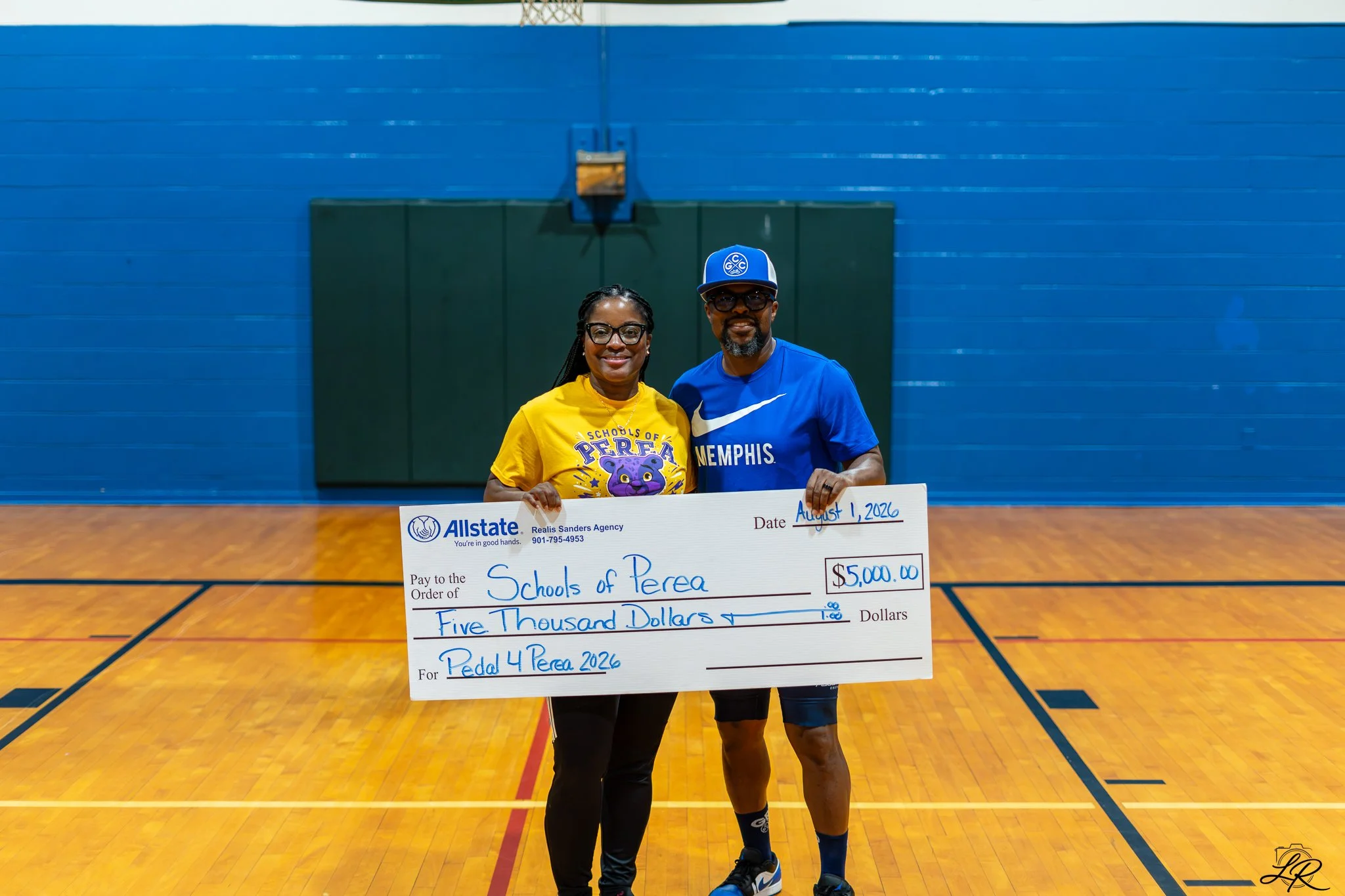 Two people standing in a gymnasium, holding a large check for $5,000. The woman on the left is wearing glasses and a yellow shirt with purple lettering, and the man on the right is wearing glasses, a blue cap, and a blue shirt. They are smiling at th
