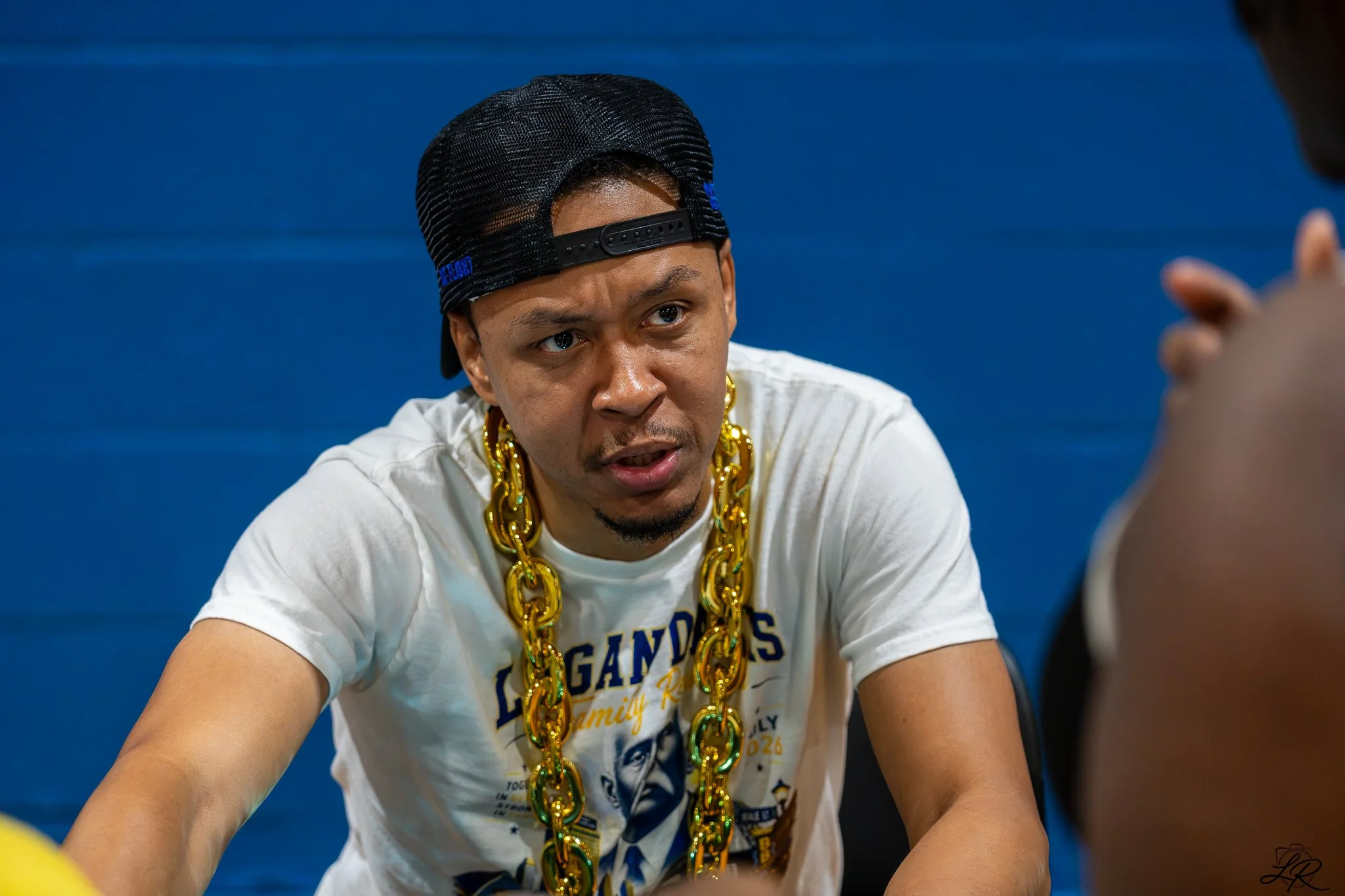 A man wearing a black baseball cap backward, a white T-shirt with Chicago themes, and large gold chains around his neck is looking at someone off-camera.