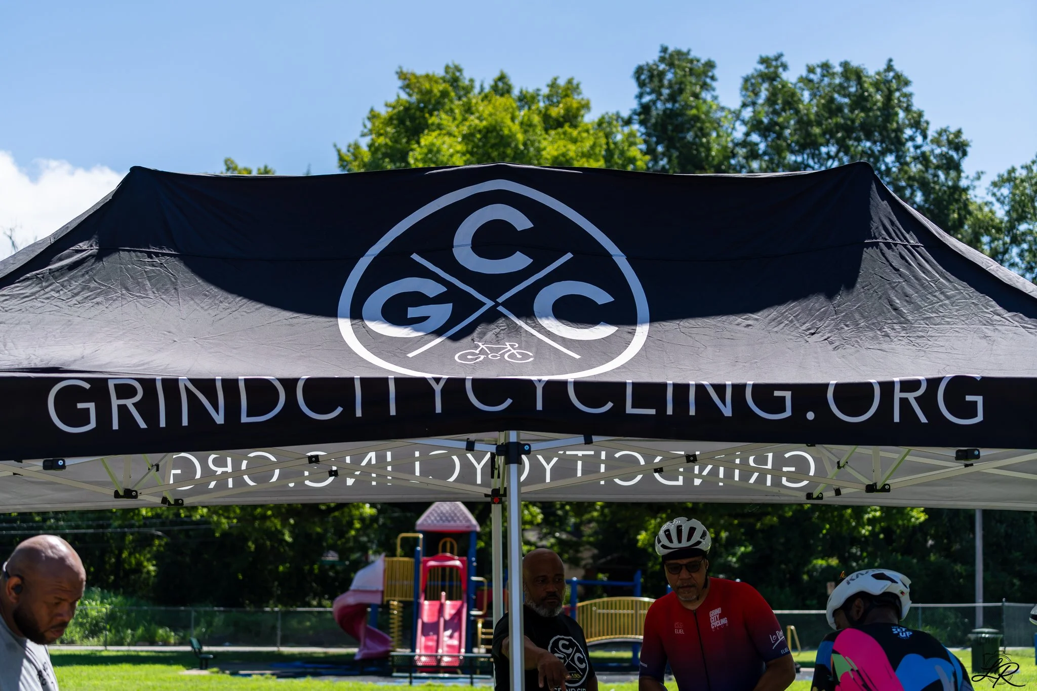 Outdoor scene with a black tent featuring a white logo for Grind City Cycling in a park with trees and play equipment, and people wearing cycling gear.