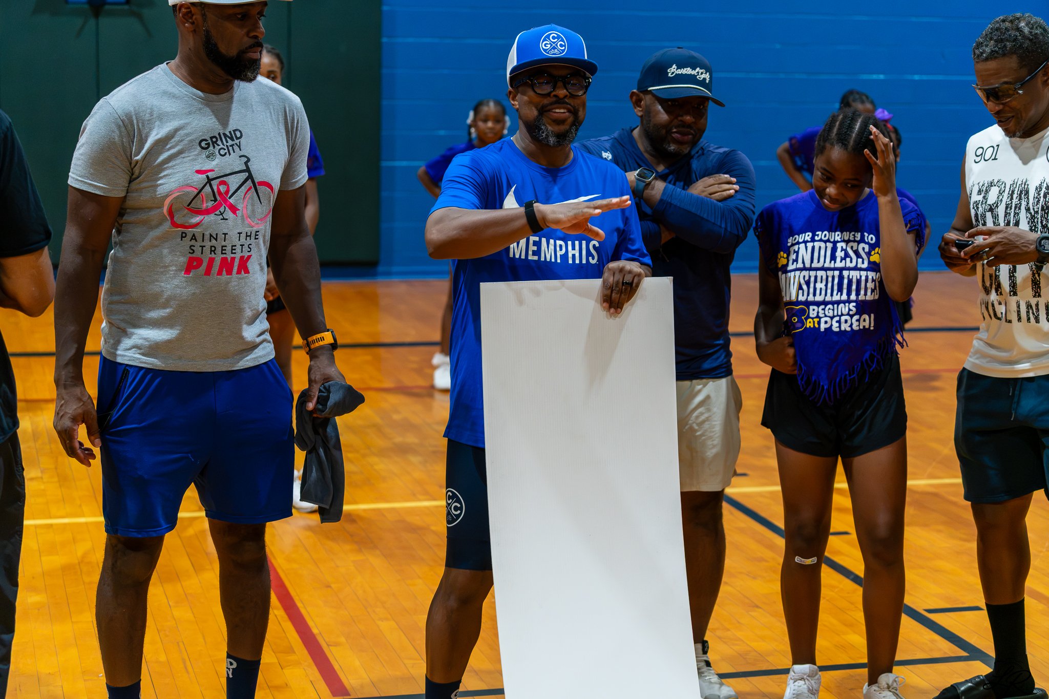 A group of people standing in a gymnasium, with one person in the center gesturing with his right hand. The group includes men and young girls, some wearing blue shirts and caps. One man is wearing a gray shirt with a bicycle graphic and the text 'GR