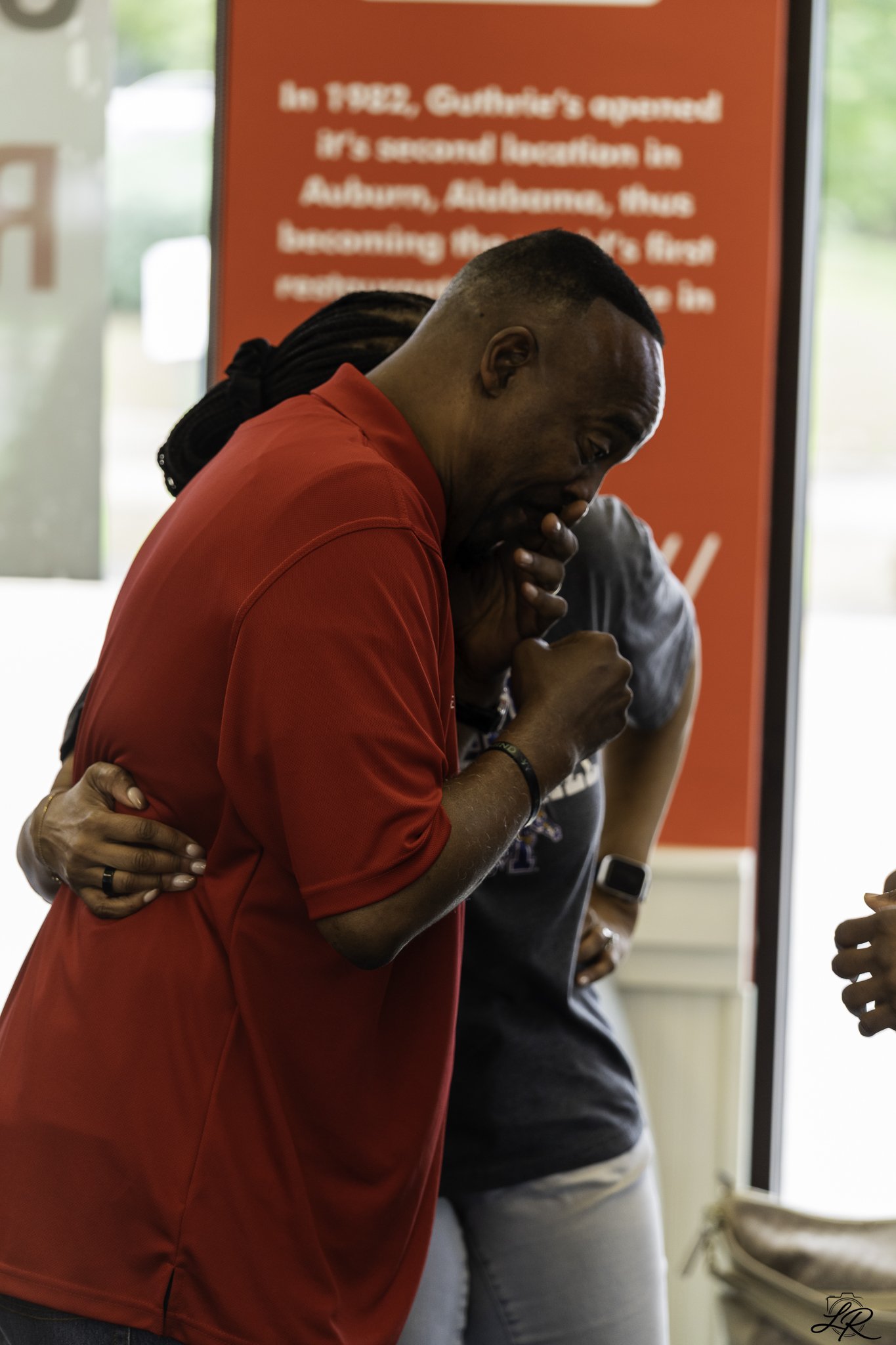 A man in a red shirt is smiling and being embraced by someone with a hand on his shoulder. He has braided hair and is wearing a black bracelet and a ring. The background shows a red sign with blurred white text.