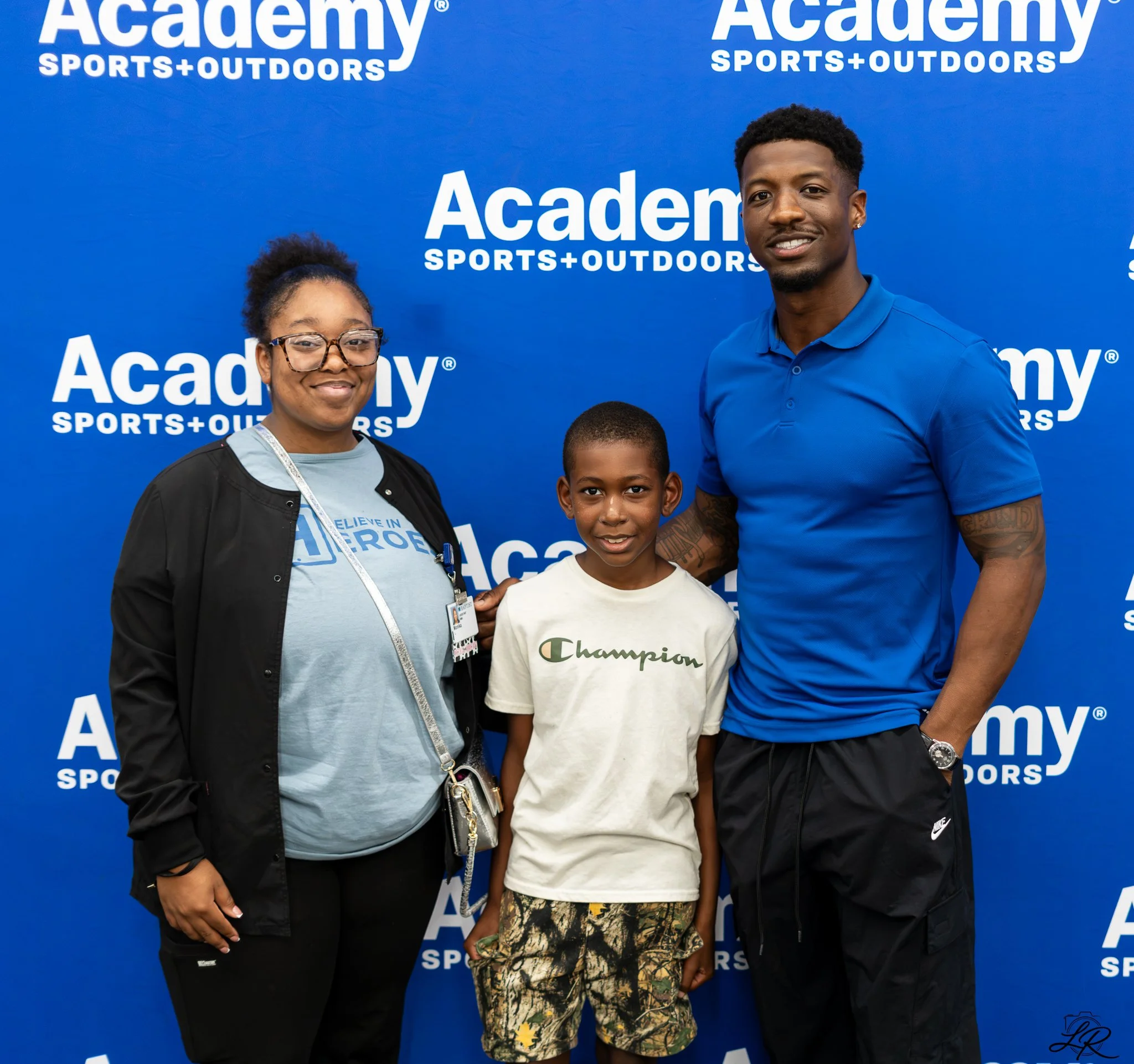 Group of three people smiling in front of a blue background with white text reading "Academy Sports + Outdoors"; a woman wearing glasses and a black jacket, a young boy in camouflage shorts, and a man with tattoos on his arm wearing a blue polo shirt